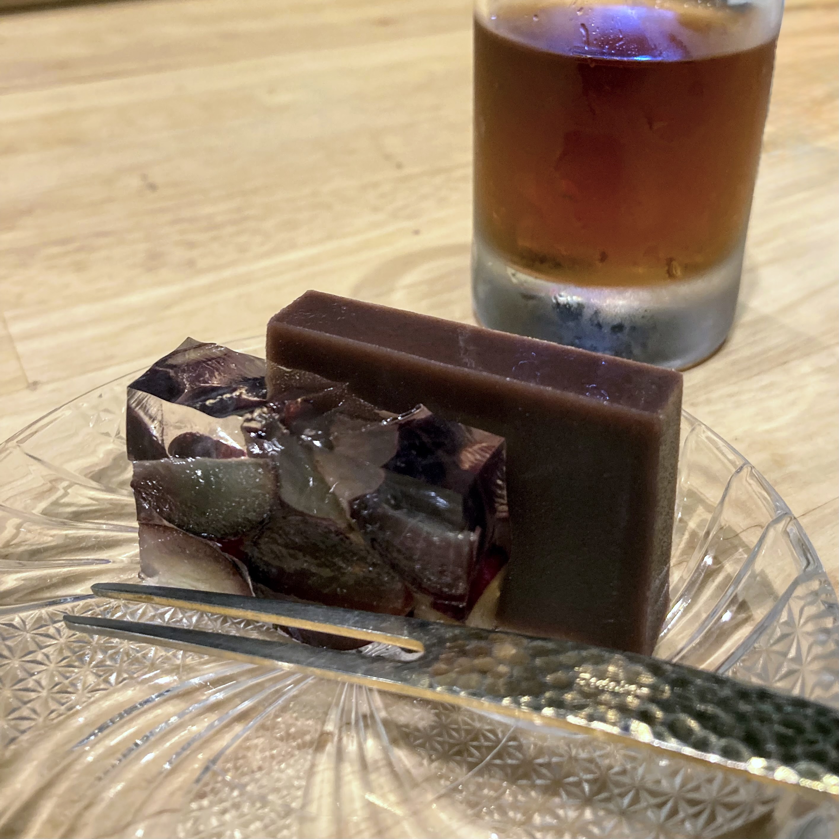 Two blocks of dark red yokan on a crystal glass plate alongside a glass of cold tea, with tweezers and a ceramic piece in the foreground.