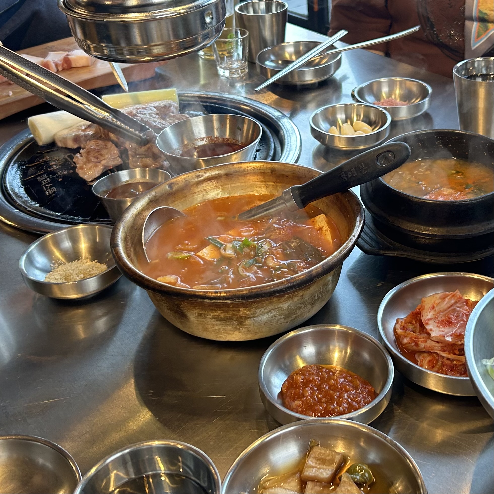 A Korean BBQ table with a bubbling jjigae in a worn aluminium pot at the center, surrounded by small banchan bowls, kimchi, and raw meat on the grill.