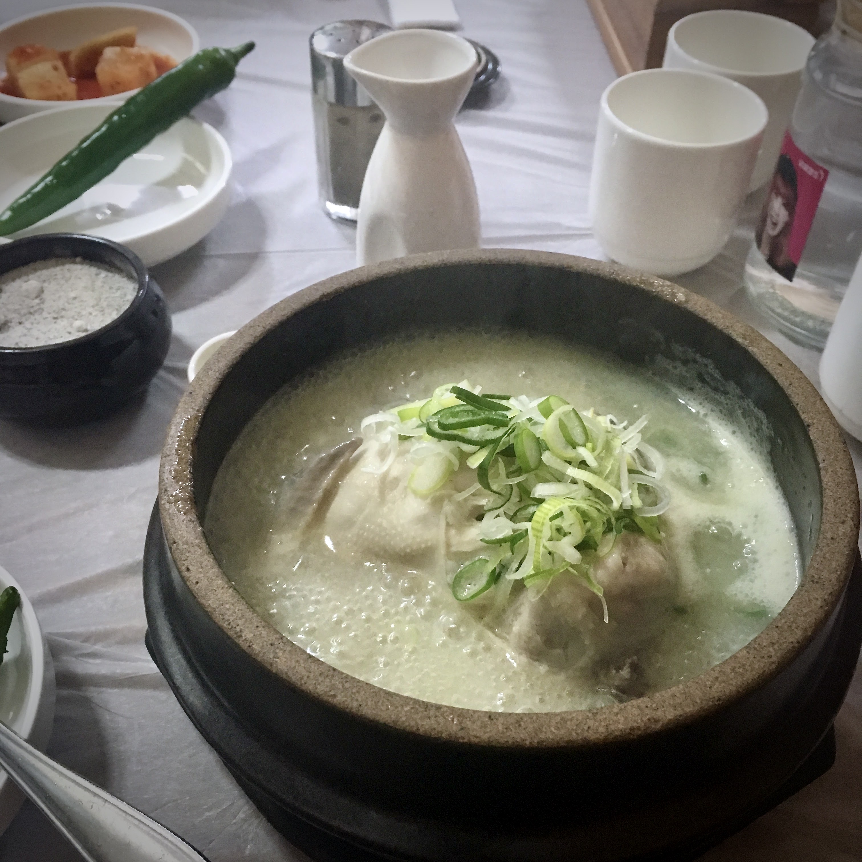 A whole samgyetang still bubbling in a dark stone ttukbaegi, topped with sliced green onion, with a green chili, salt bowl.