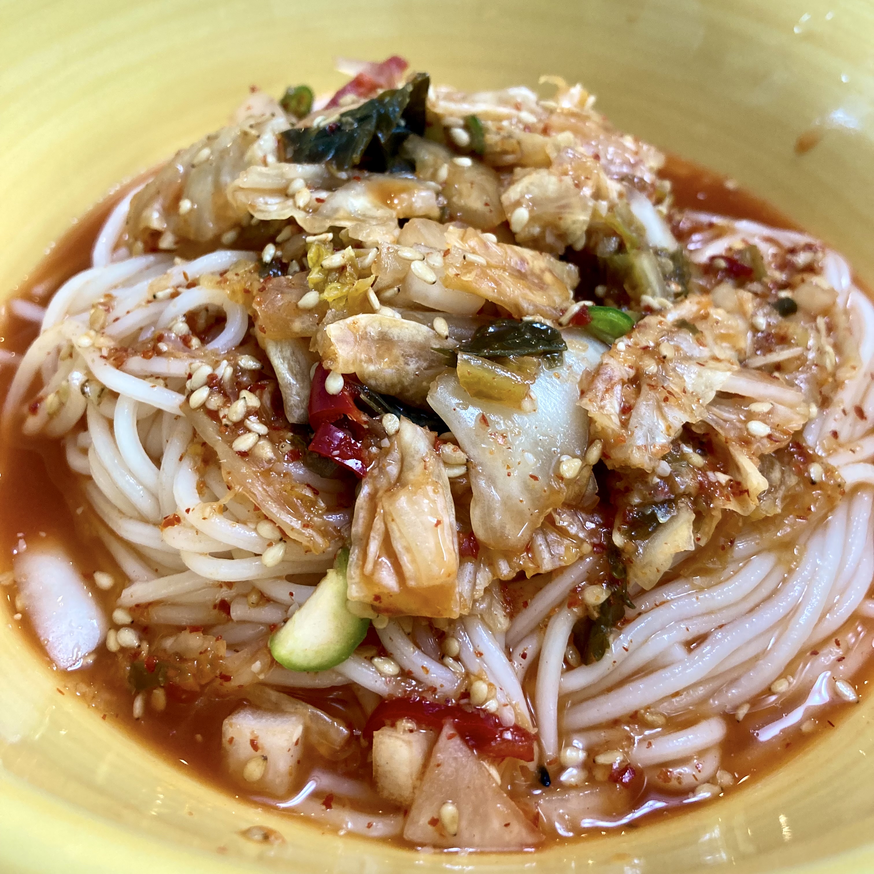 A yellow bowl of kimchi-mari guksu — thin white noodles in red kimchi brine, topped with aged kimchi, cucumber, sesame seeds, and gochugaru.