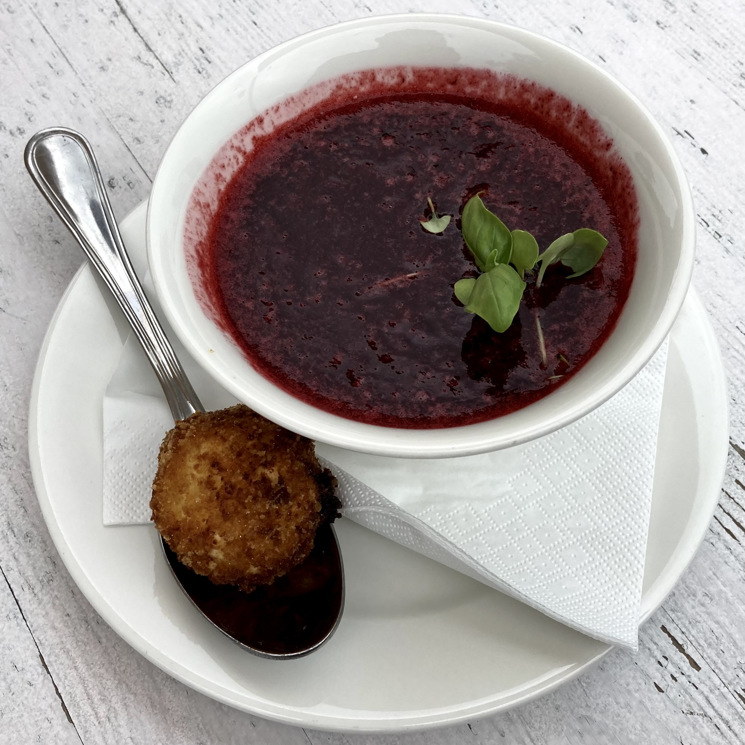 A white bowl of deep red Hungarian cold sour cherry soup garnished with fresh basil, served with a fried ball on a spoon alongside.