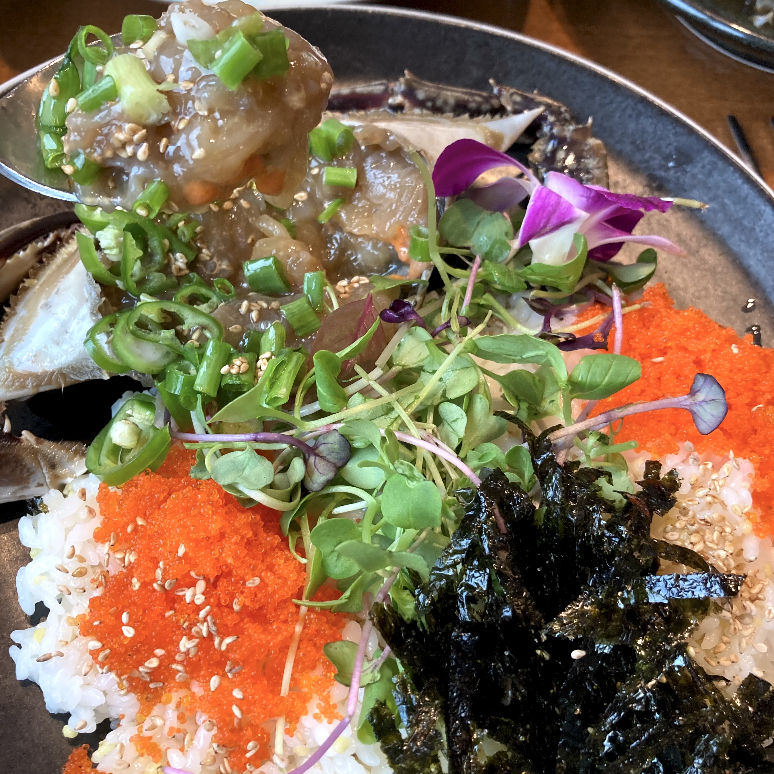 A dark plate with rice topped with scraped ganjang gejang meat, bright orange fish roe, crumbled gim, microgreens, sliced green onion, and edible flowers, with crab shell visible at the edge.