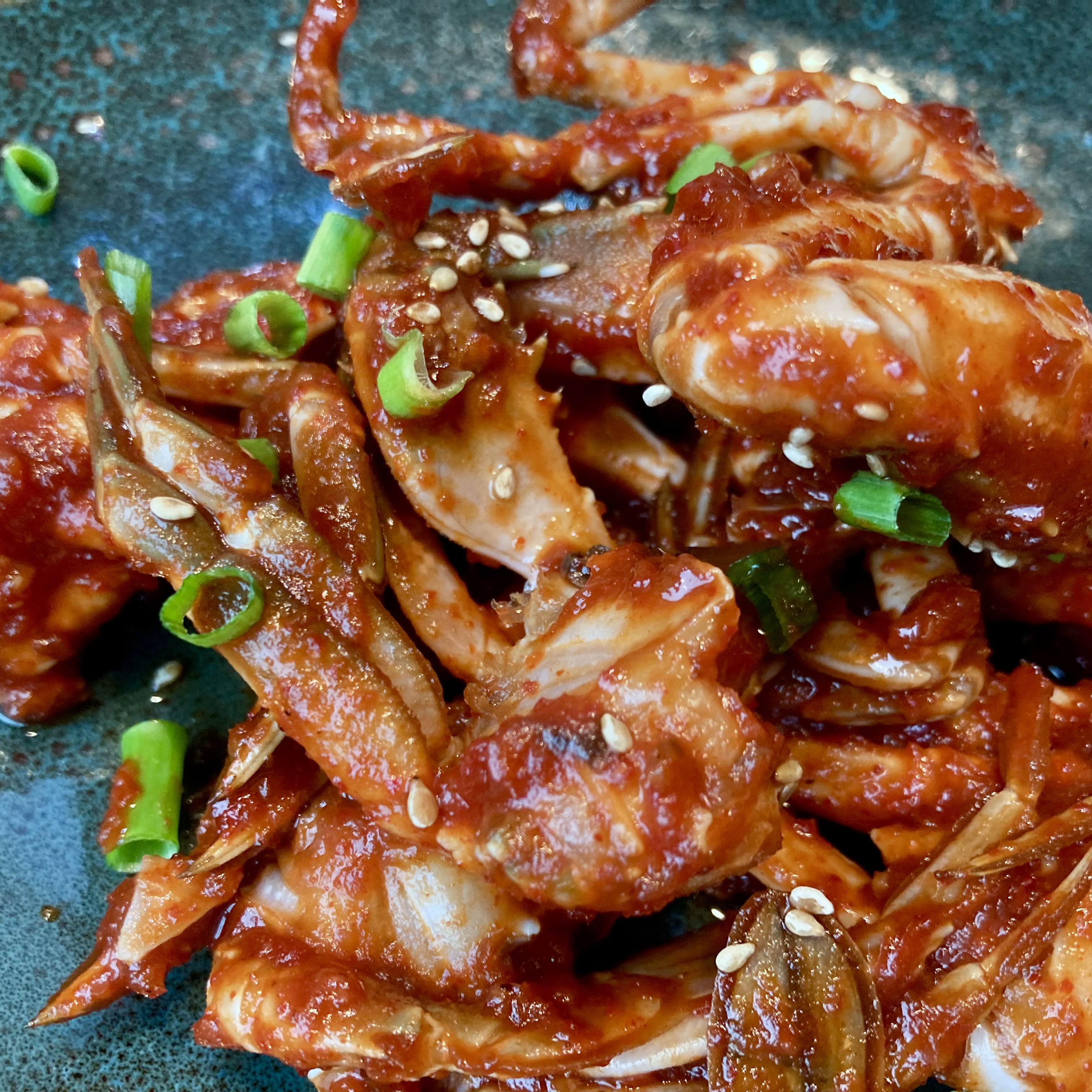 A dark plate piled with yangnyeom gejang, crab pieces heavily coated in red gochujang sauce, topped with sliced green onion and sesame seeds.