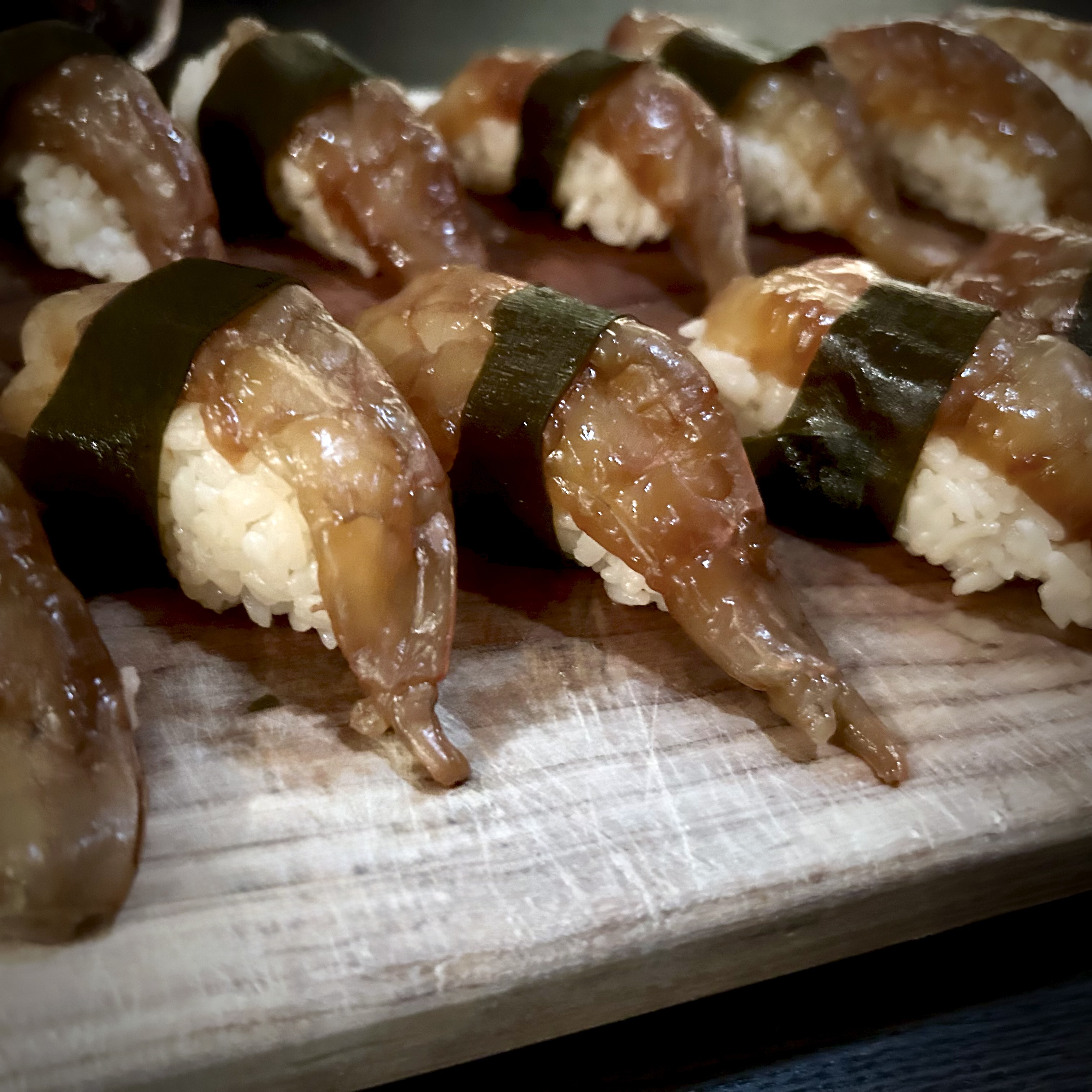 Several pieces of ganjang saewoo chobap on a wooden board, each marinated shrimp pressed over a small rice mound and wrapped in a strip of dasima.