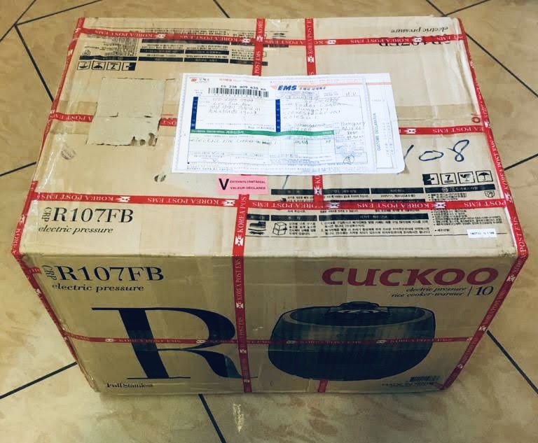Cardboard box printed with "Cuckoo electric pressure rice cooker warmer," wrapped in Korea Post EMS tape with a shipping label and customs form attached, sitting on a tiled floor.