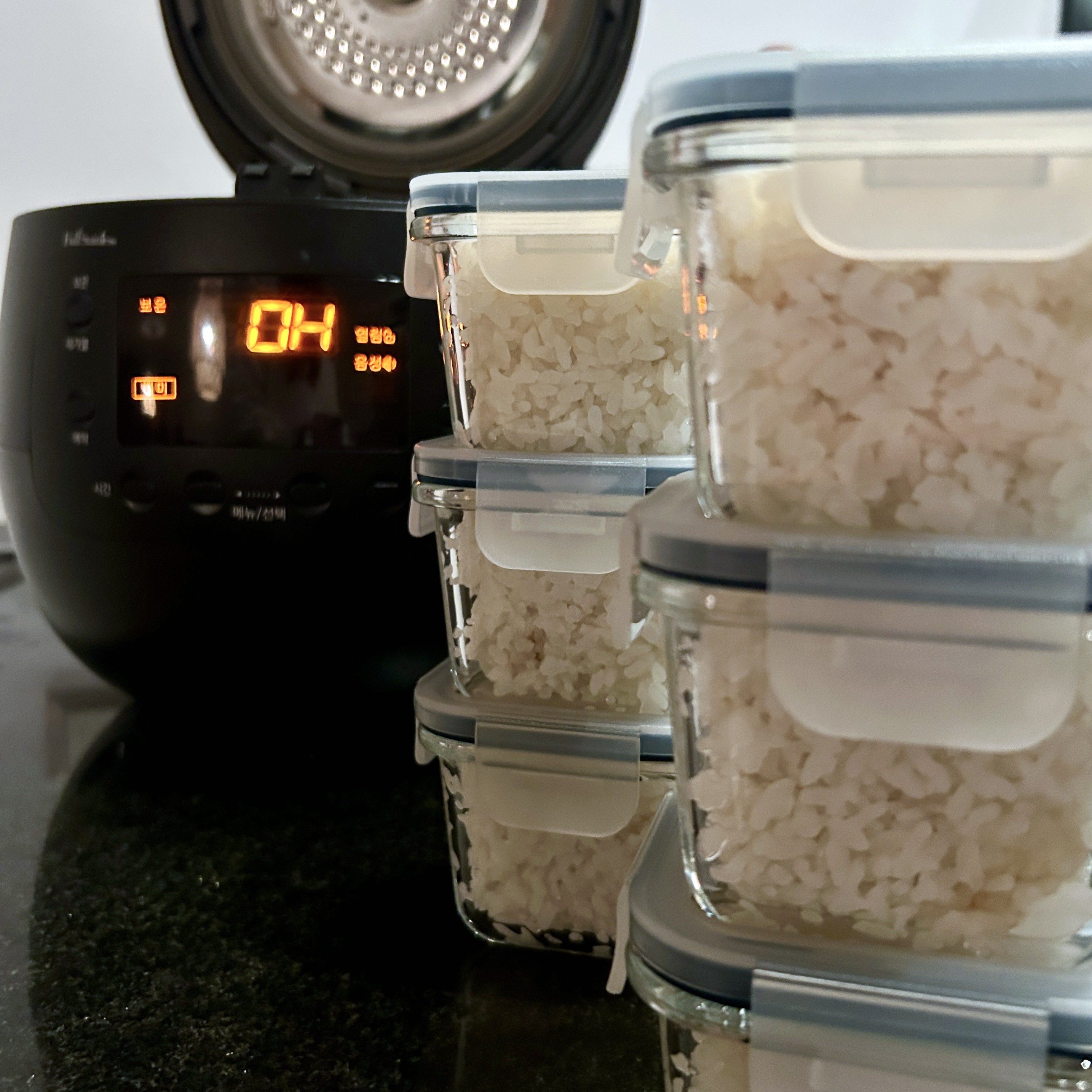 Black rice cooker with lid open and digital display lit, beside stacked glass containers filled with cooked rice, ready to be frozen.