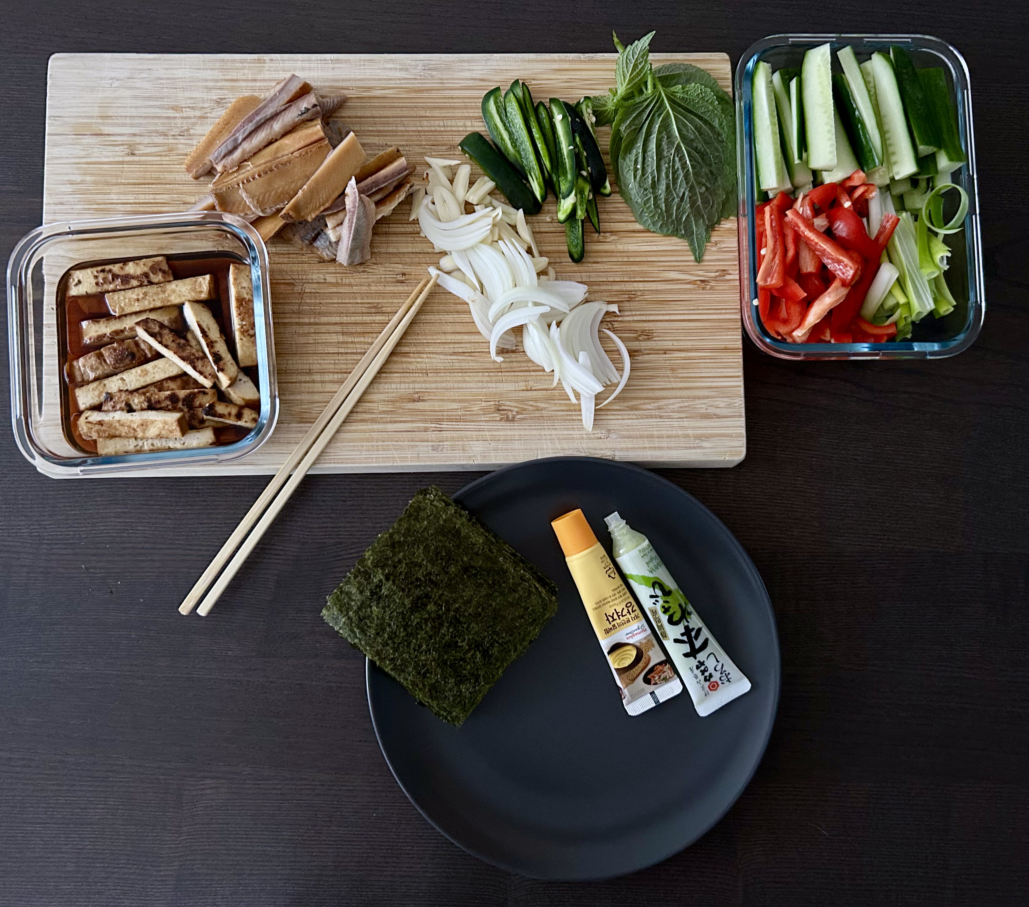 temaki ingredients laid out on a bamboo board and containers: marinated tofu, smoked fish, sliced onion, cucumber, kkaennip, nori sheets, wasabi and soy sauce tubes, and julienned vegetables in a glass container.