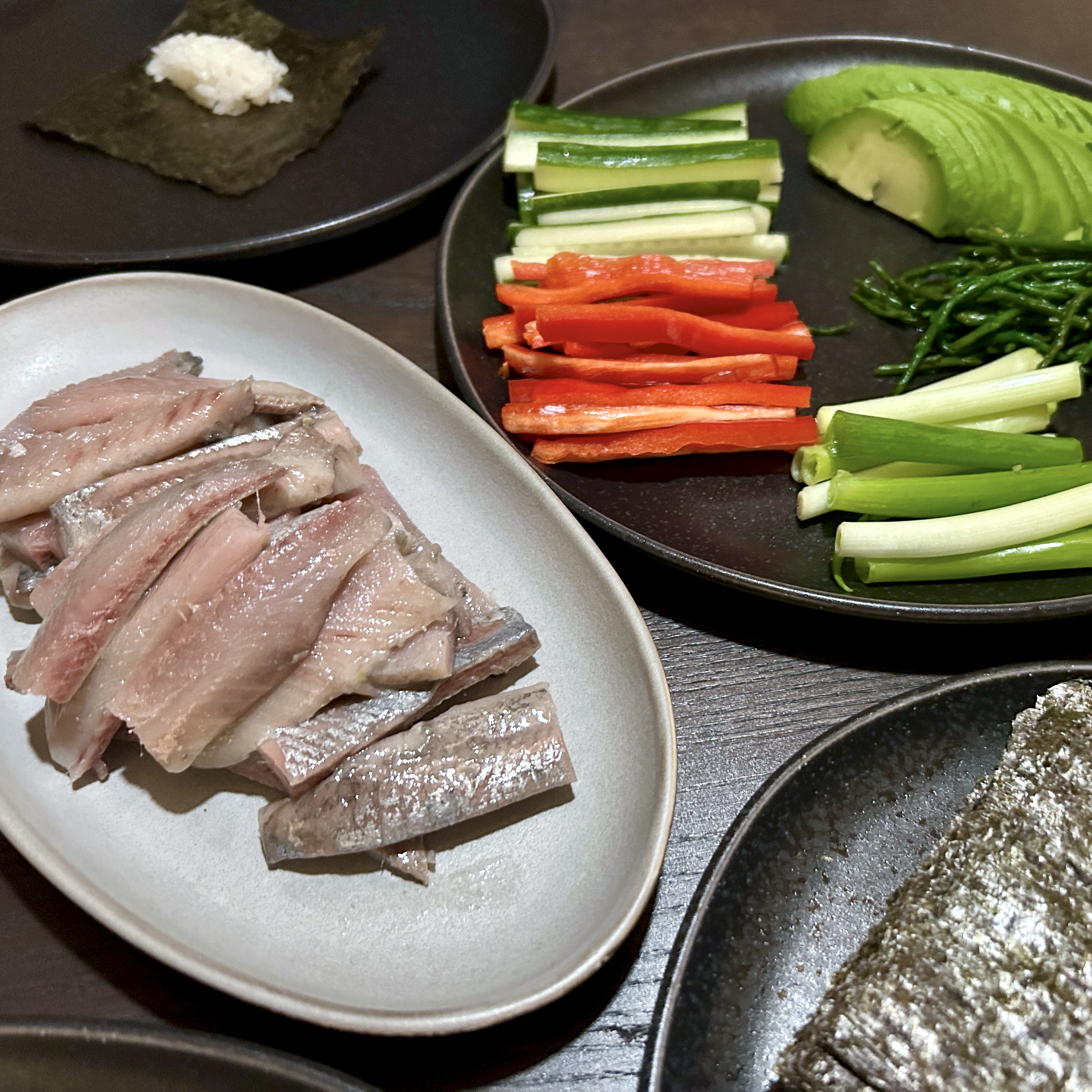 A table set for temaki with sliced nieuwe haring on a white plate, julienned vegetables and avocado on a dark plate, nori sheets, and a piece of nori with a small mound of rice.