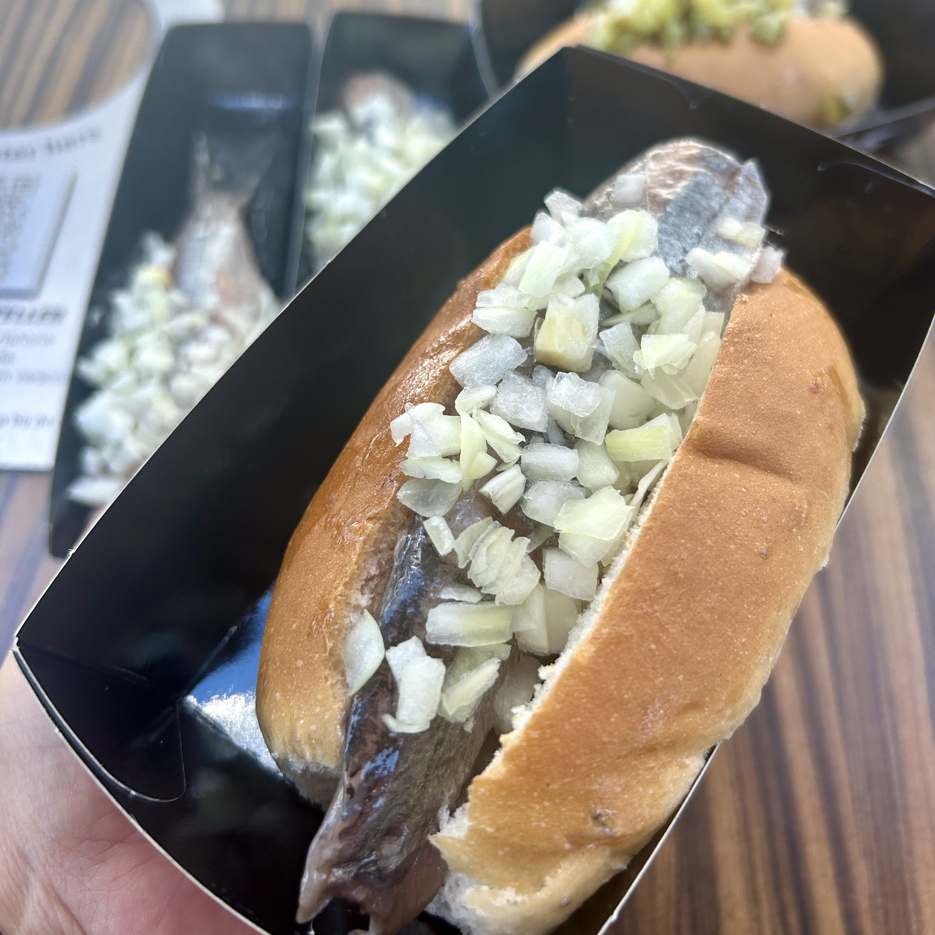 A hand holding a broodje haring in a black cardboard holder, a soft white bun filled with nieuwe haring and topped with chopped raw onion.