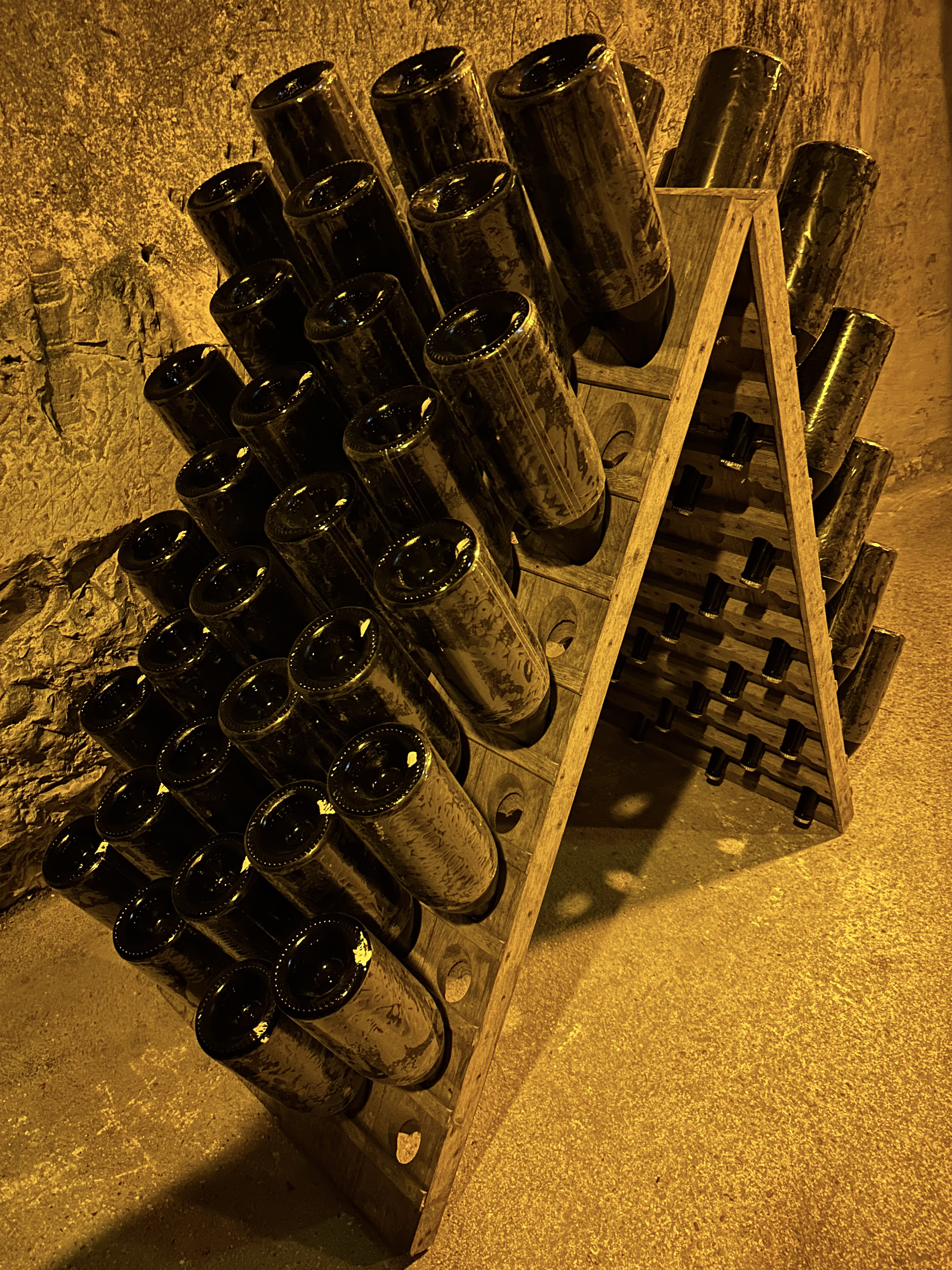 A large wooden A-frame rack holding rows of dark champagne bottles resting neck-down in a stone cellar, lit in amber light.