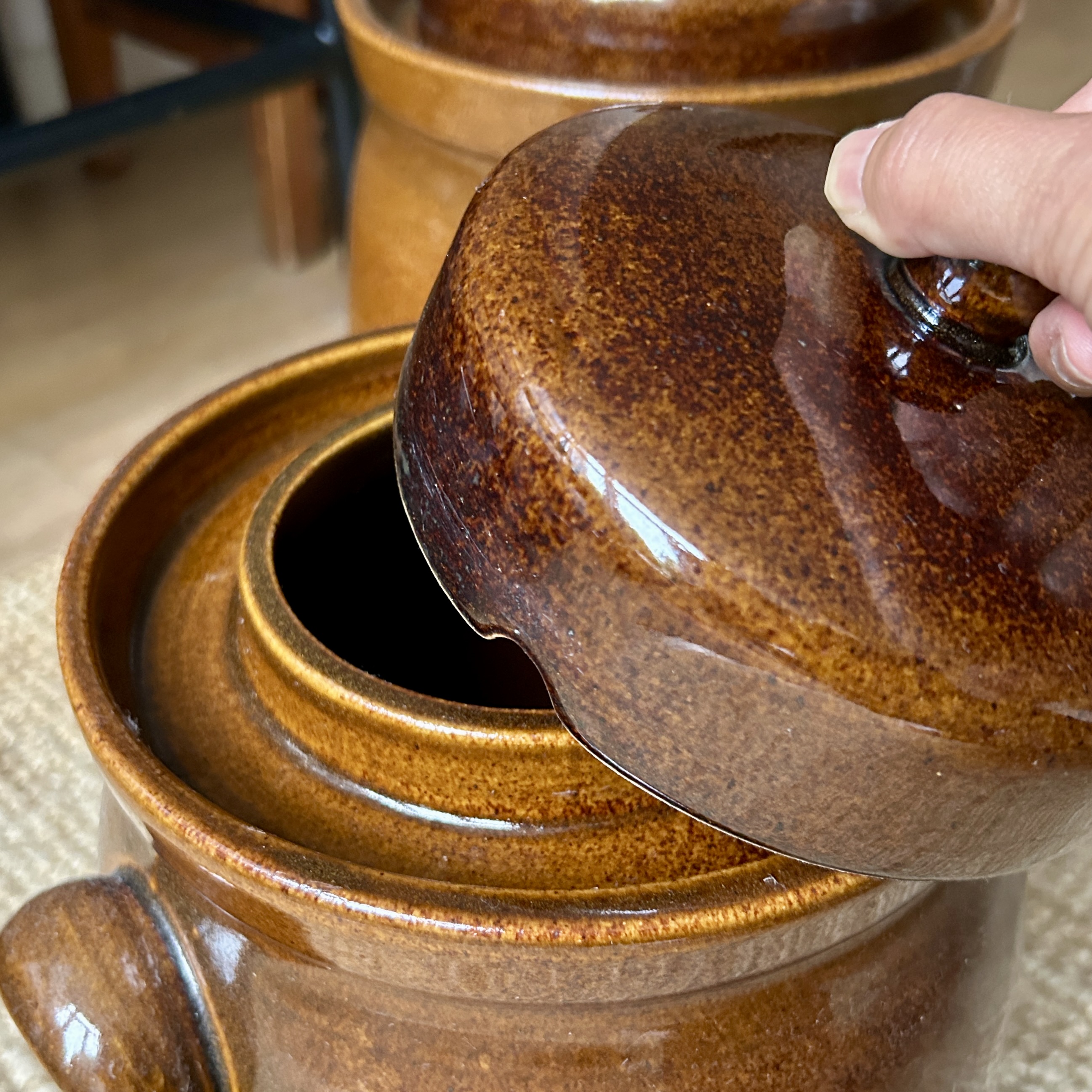 A hand lifting the domed lid off a glazed brown ceramic crock, revealing the raised inner rim and water channel around its opening.
