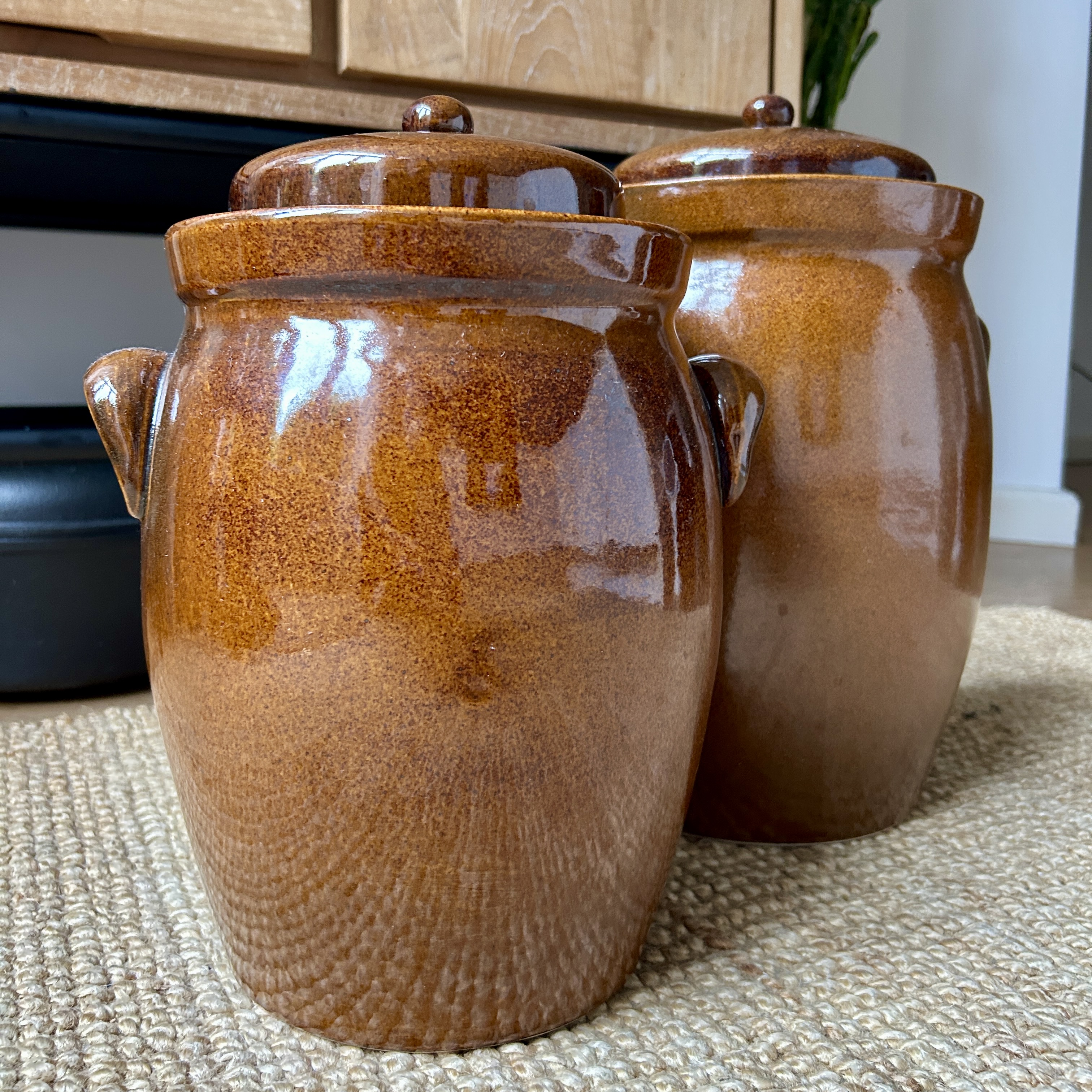 Two glazed brown ceramic crocks with domed lids and side handles, sitting on a woven jute rug in a living room.
