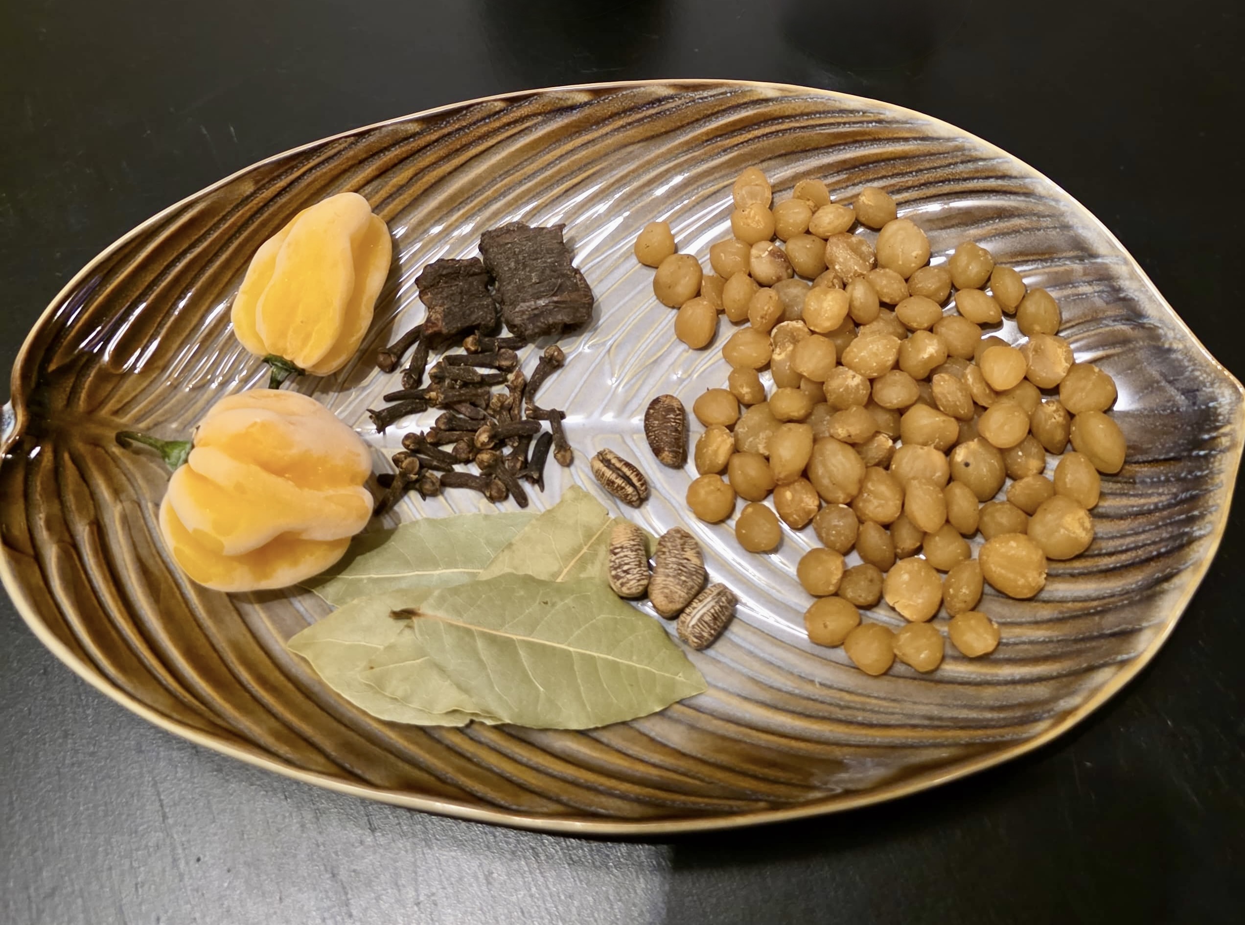A plate laid out with pepper soup ingredients: njansang seeds, dried orange peppers, bark, cloves, bay leaves, and small dark pods.
