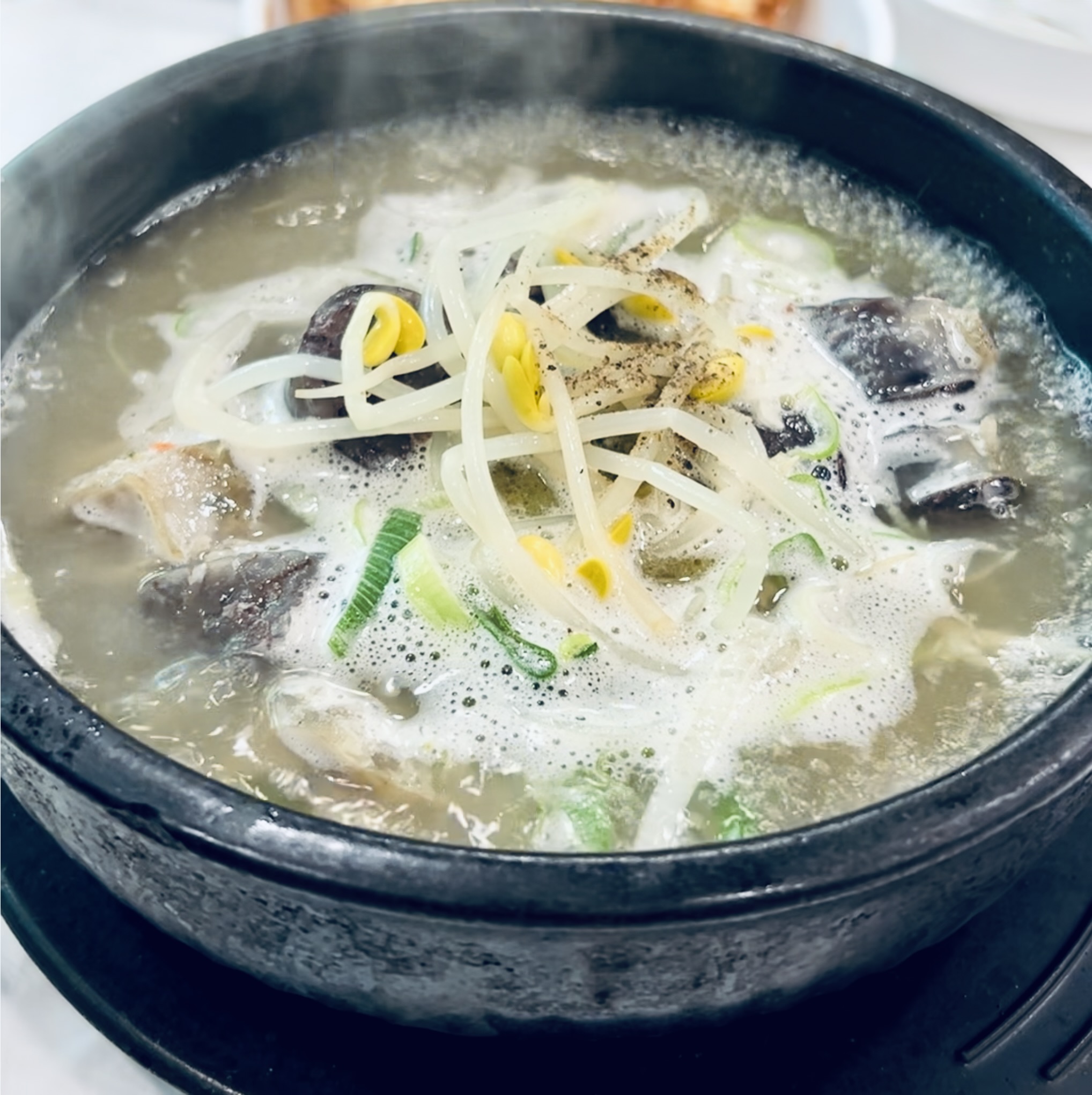 A stone bowl of milky haejangguk broth topped with bean sprouts, scallions, and cracked black pepper, steaming hot.