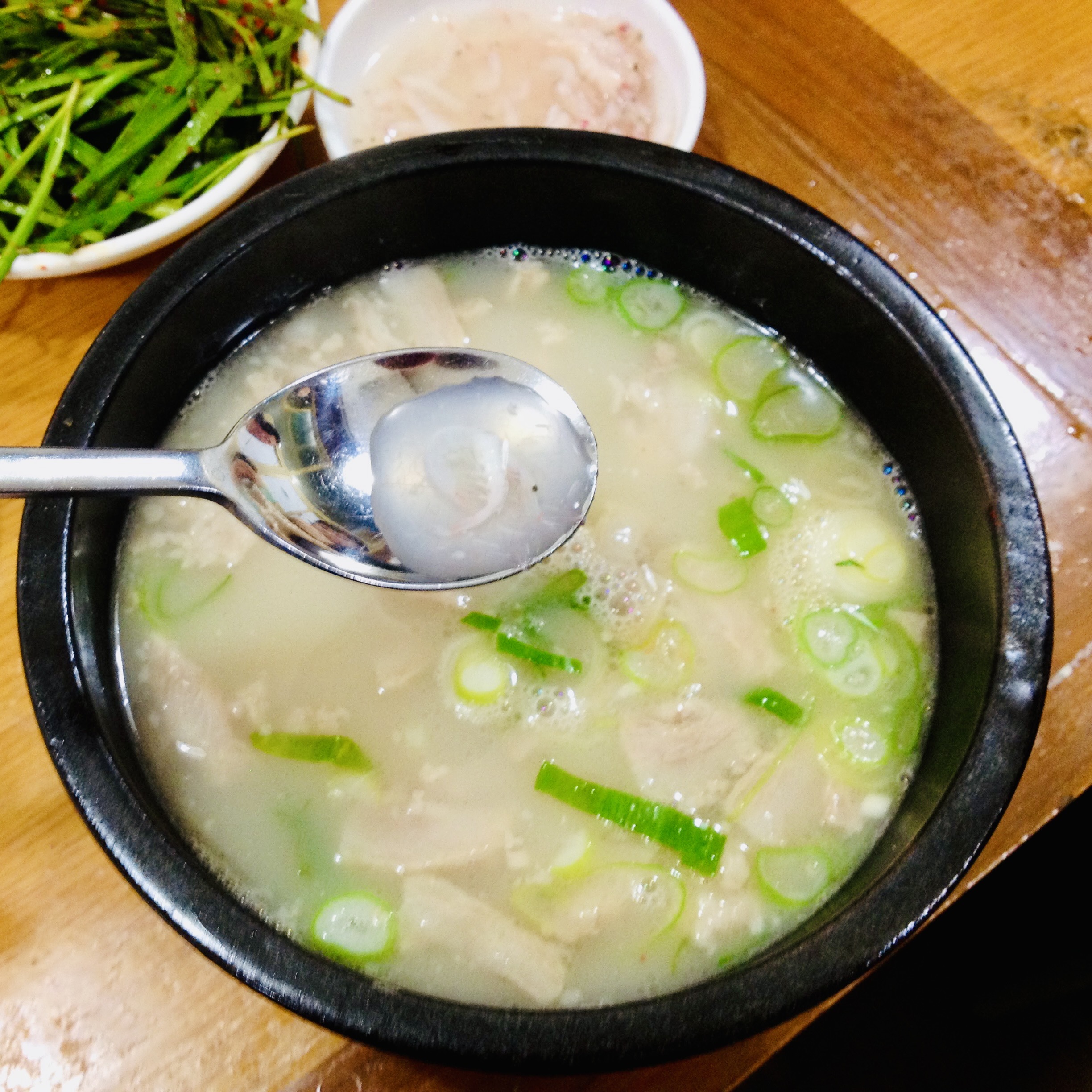A spoon lifting the first spoonful of broth from a stone bowl of soup, milky and topped with sliced scallions.