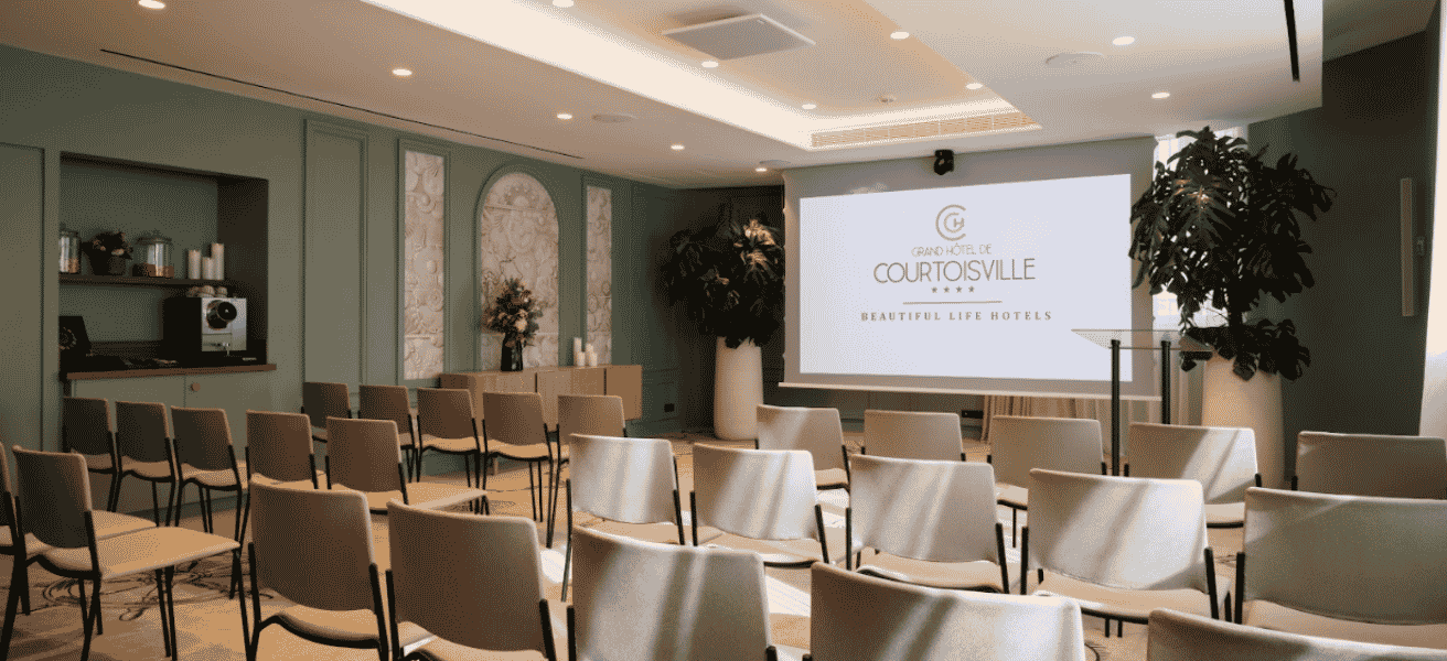 Salle de séminaire élégante avec des chaises beiges alignées face à un grand écran de projection affichant le logo du Grand Hôtel de Courtoisville.