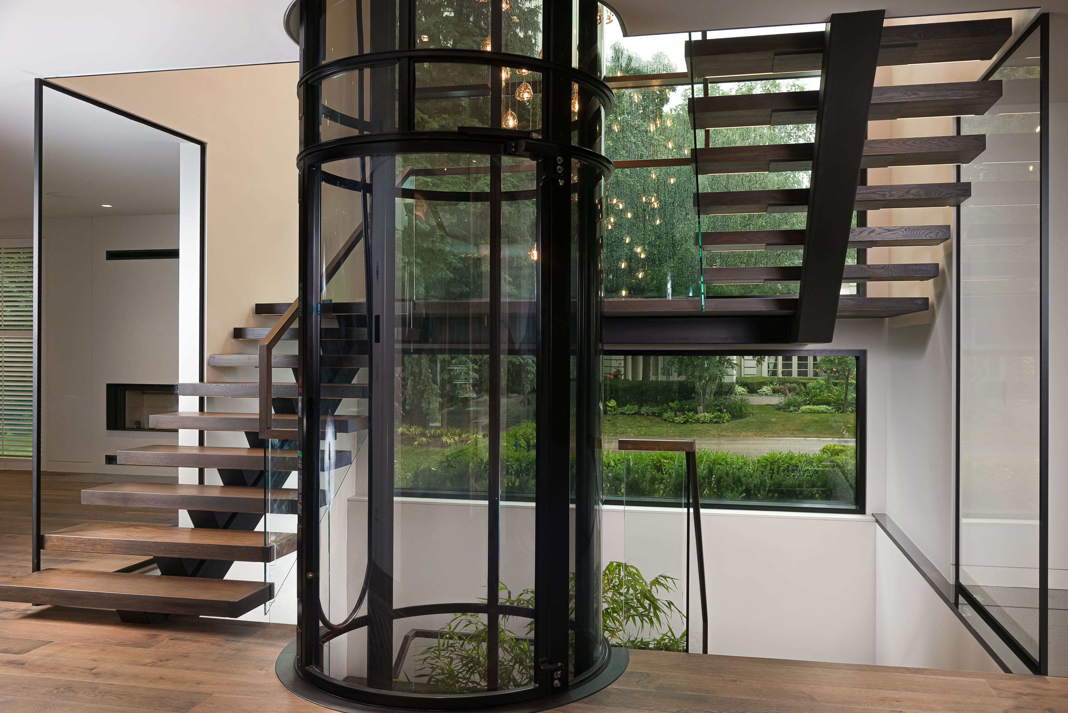 Modern interior featuring a cylindrical glass elevator and floating wooden staircase, highlighting Goosen Inc.’s innovative architectural millwork.