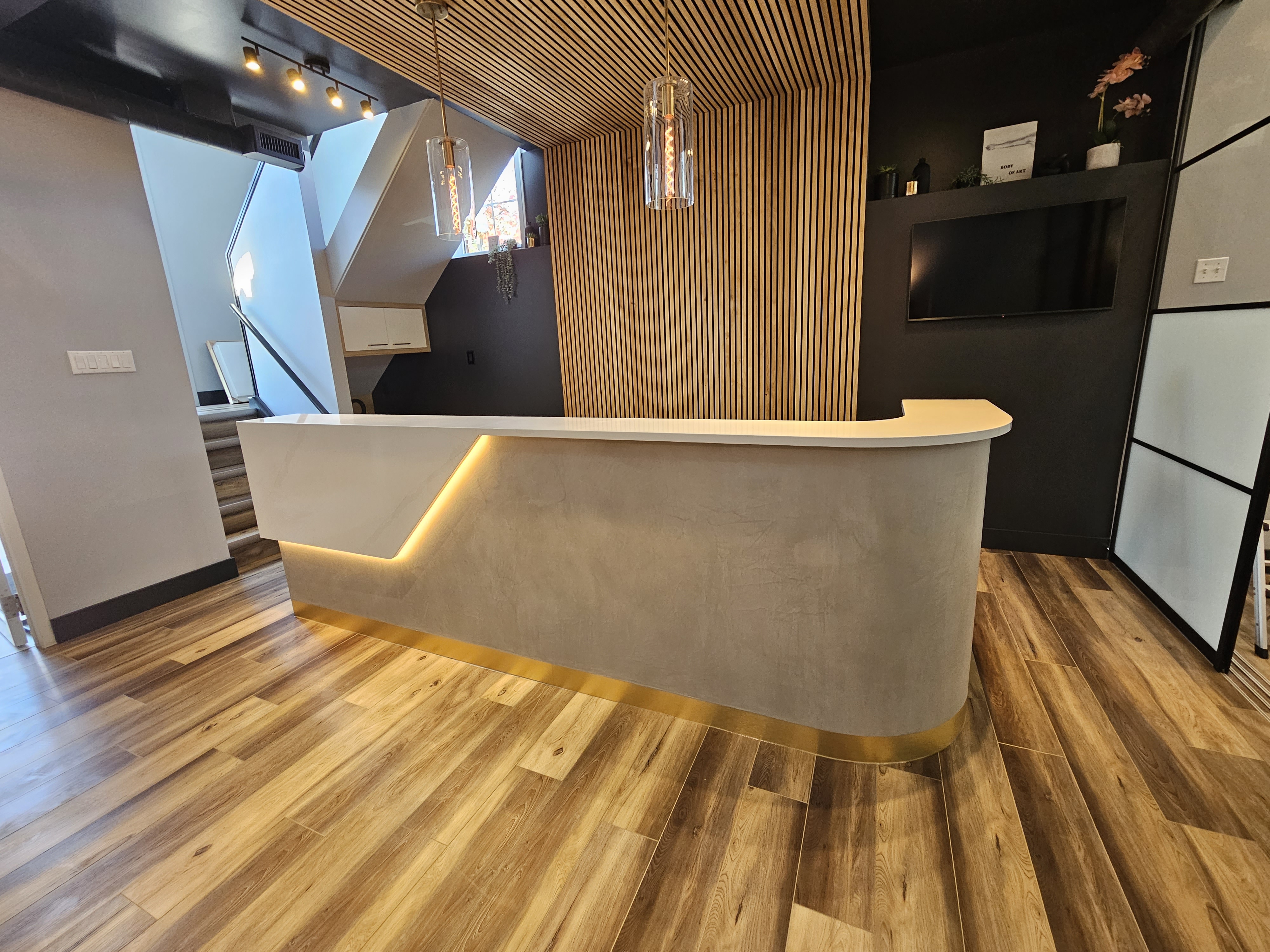 Modern reception desk with white top, beige base, and warm underlighting, set against vertical wooden slats and dark walls with a mounted TV.