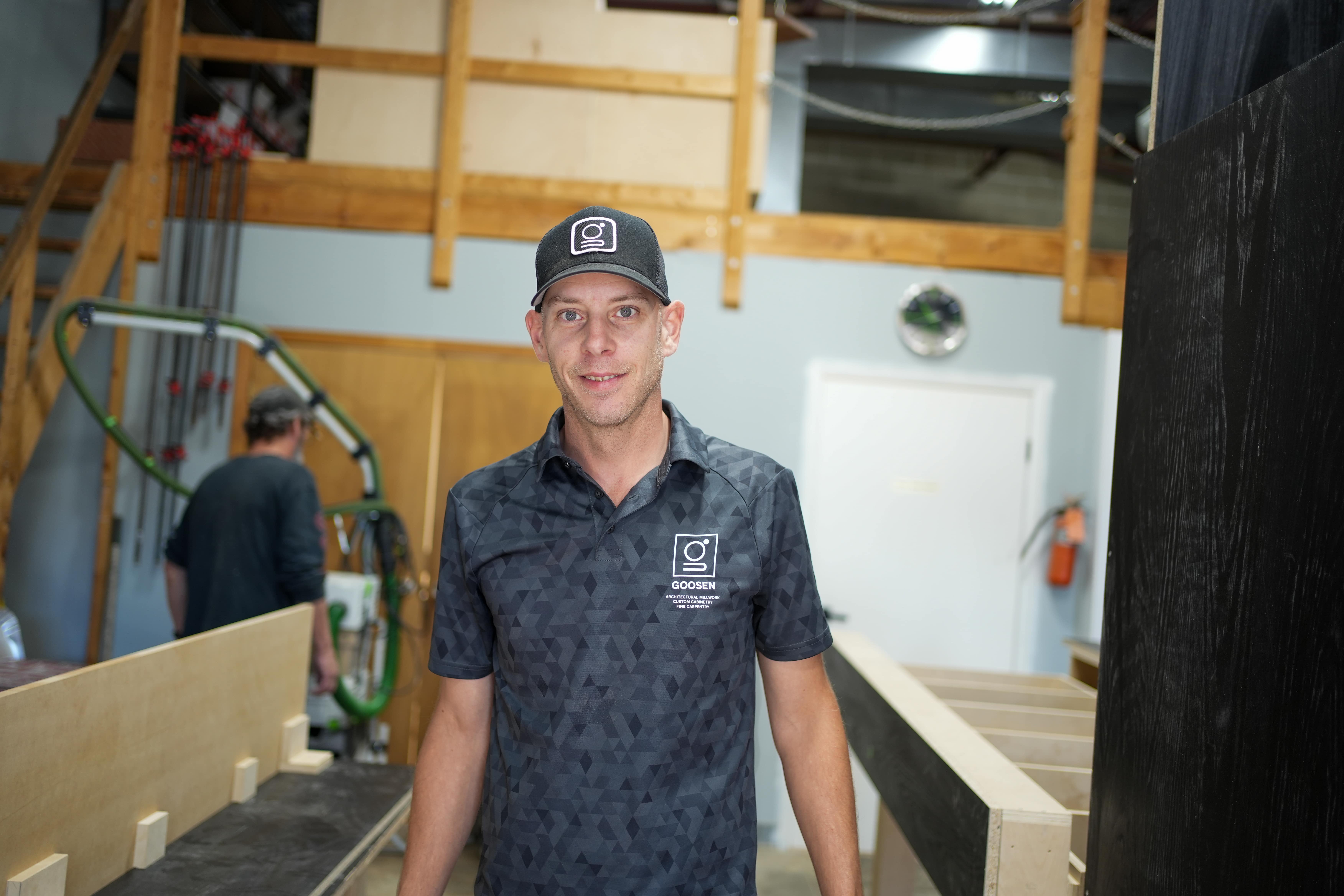 Goosen Inc. owner standing in a woodworking shop, wearing a branded shirt and cap.