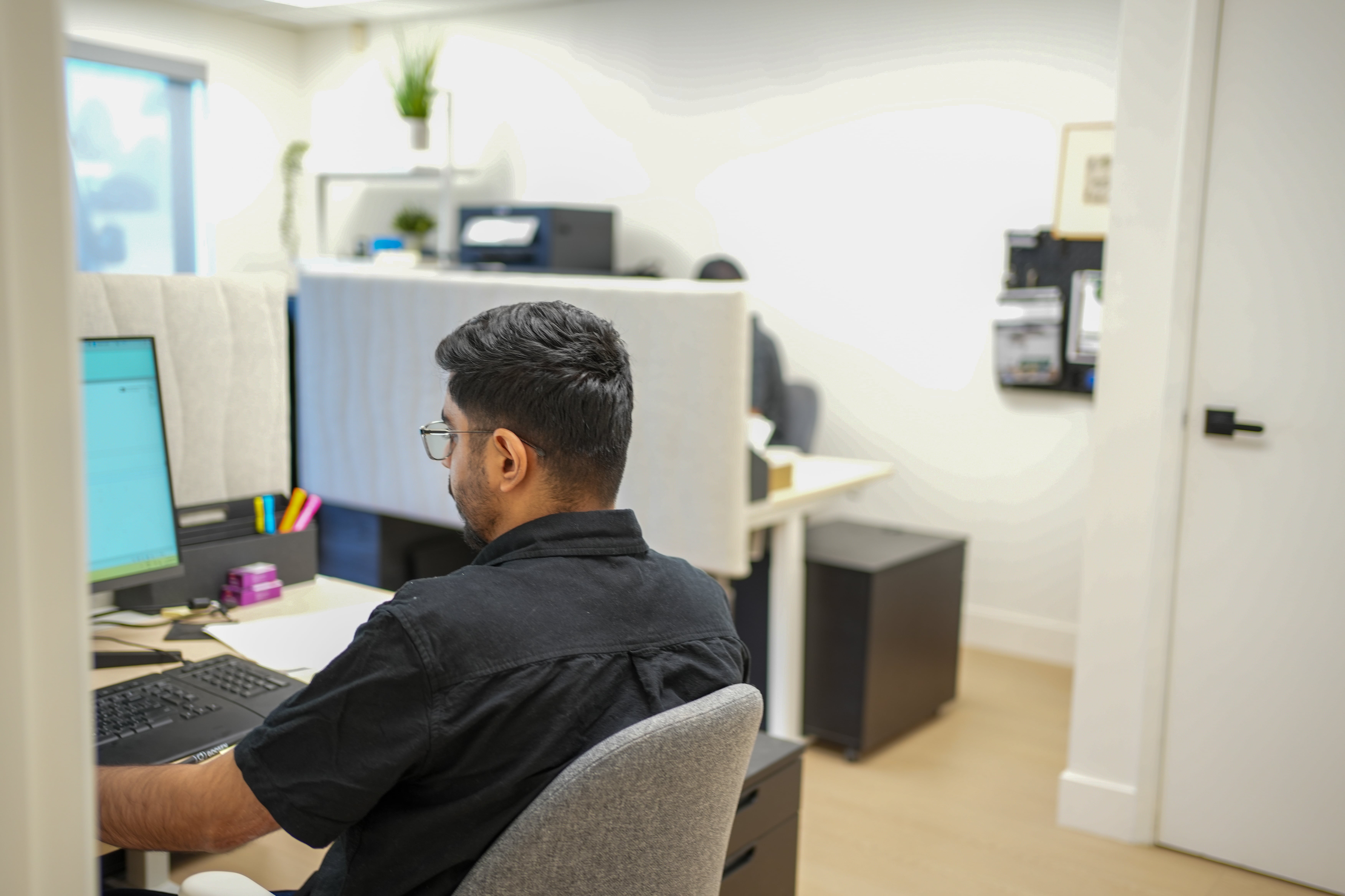 Man with glasses sitting at a desk working on a computer in a modern office space with partitioned workstations.