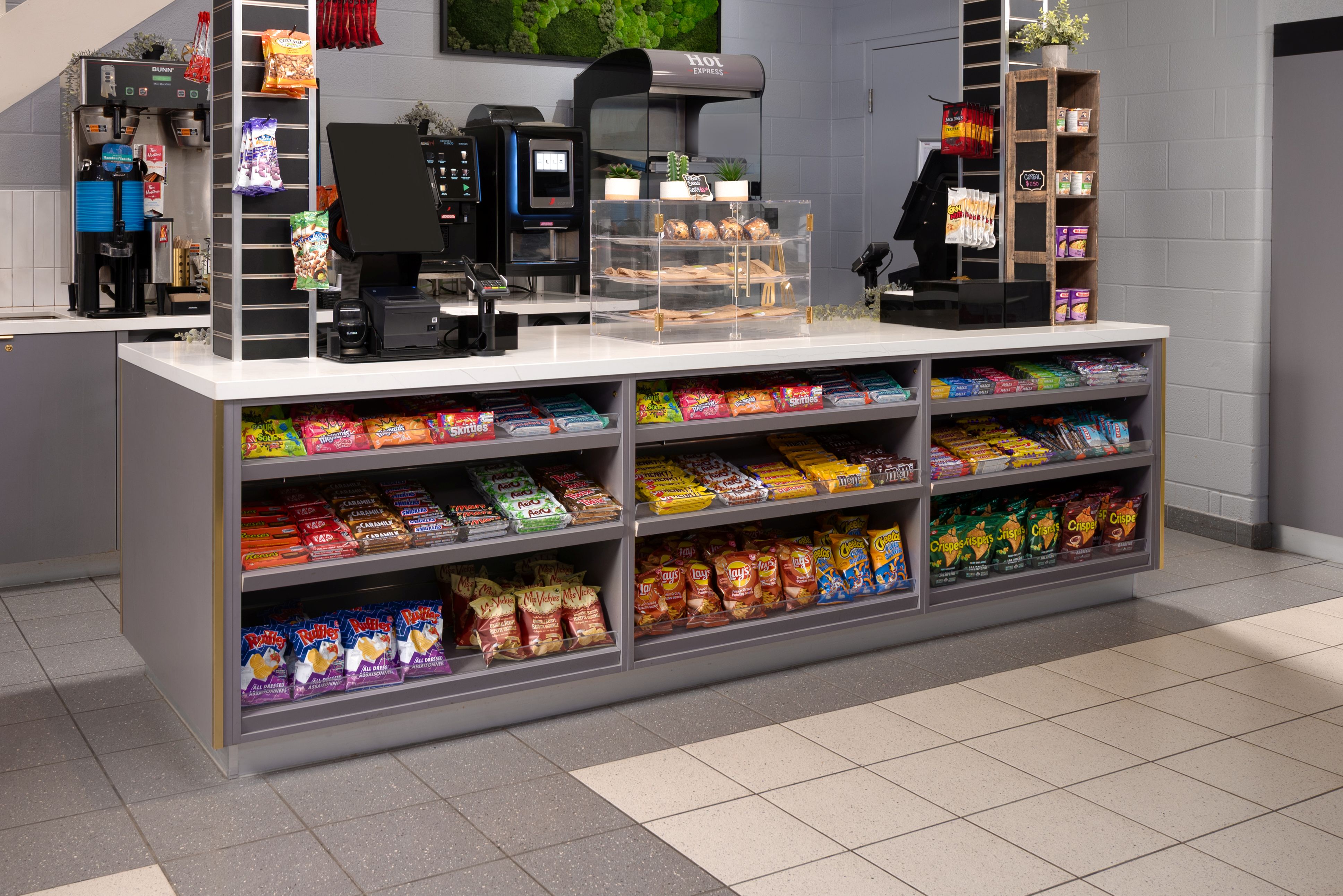 Snack counter with various chips, candy bars, and a bakery display in a café or convenience store with coffee machines in the background.