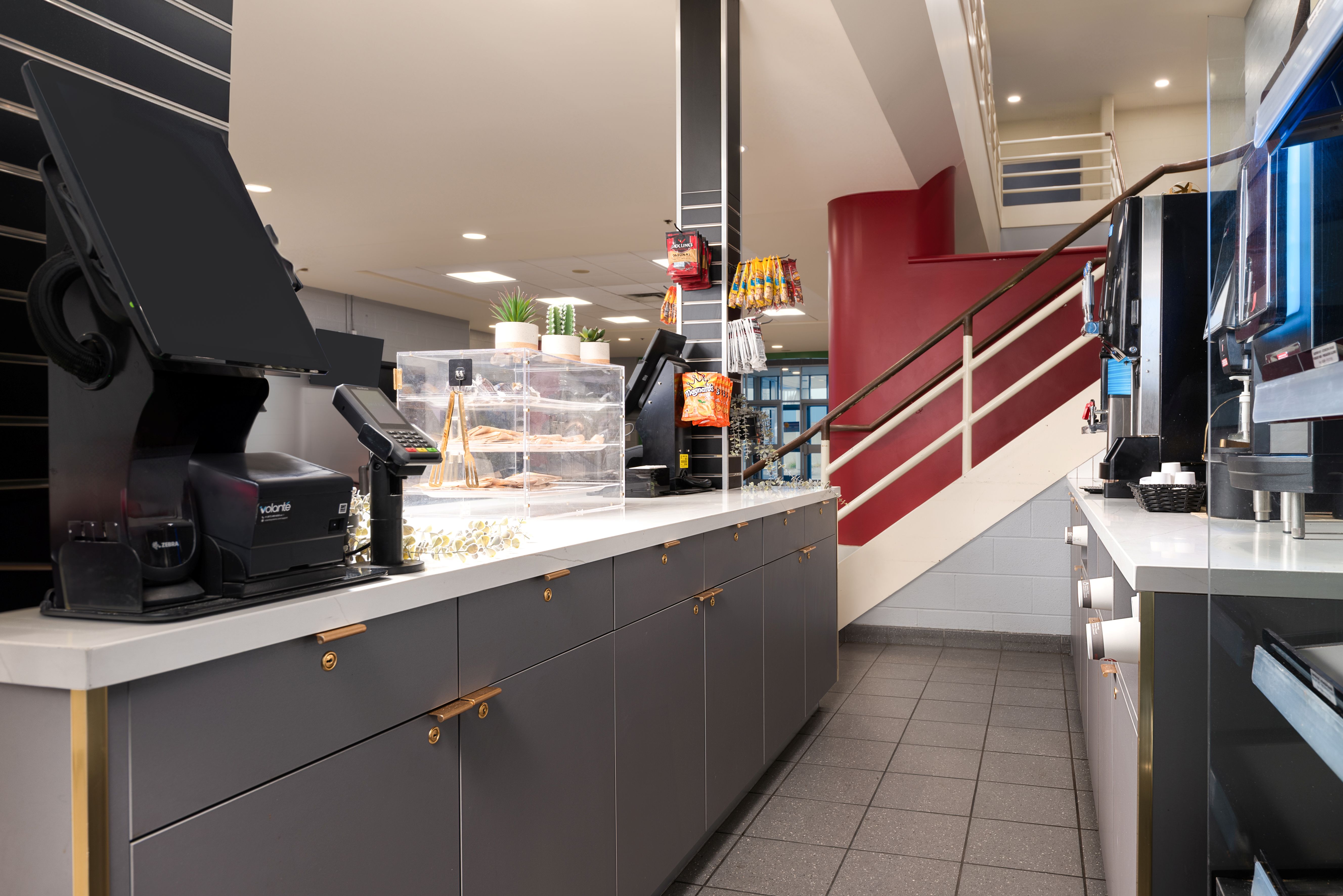 Modern cafe counter with cash registers, snack display, and coffee machines next to a red and white staircase.