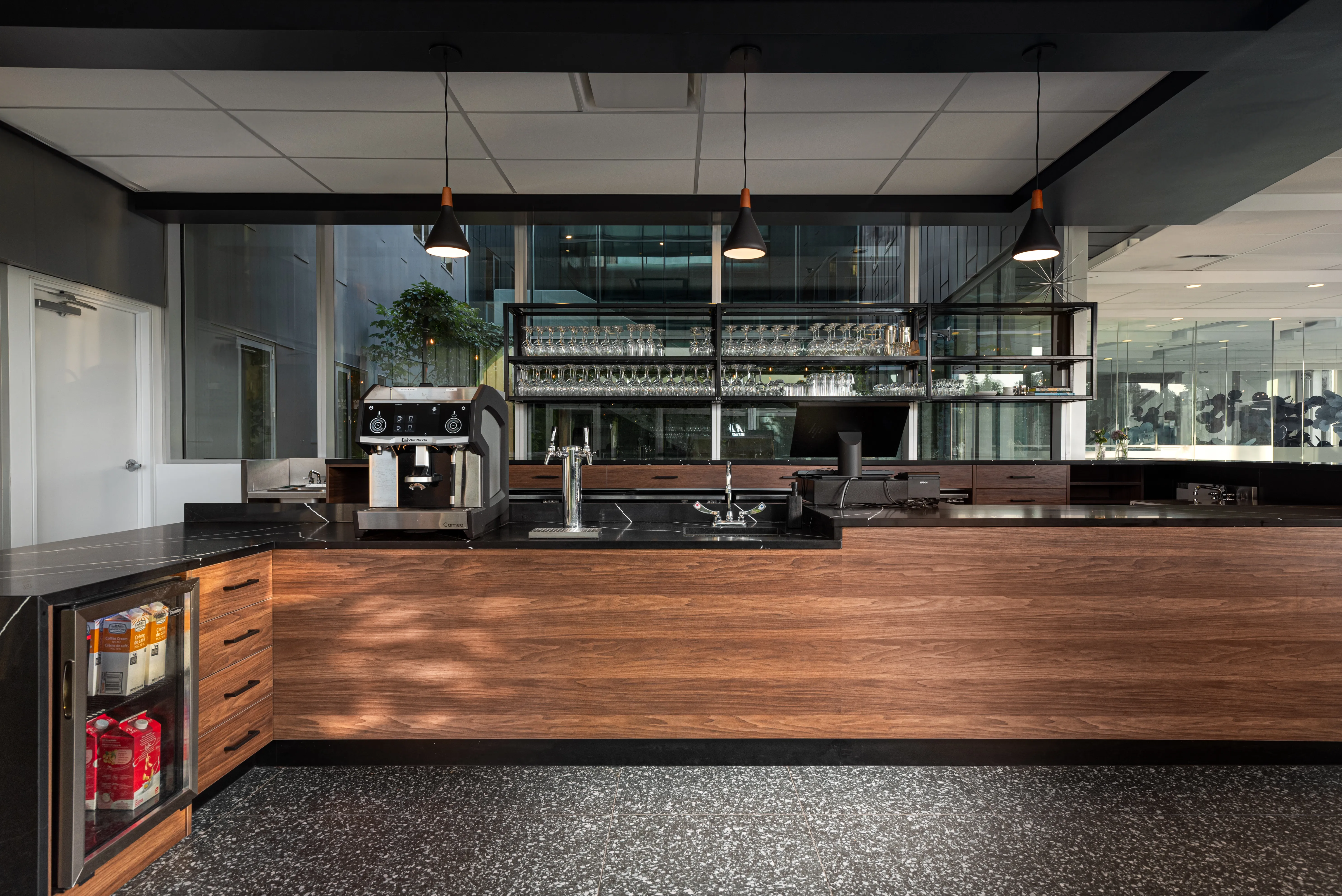 Modern coffee bar with espresso machine, glass shelves, and wooden cabinetry under three pendant lights.