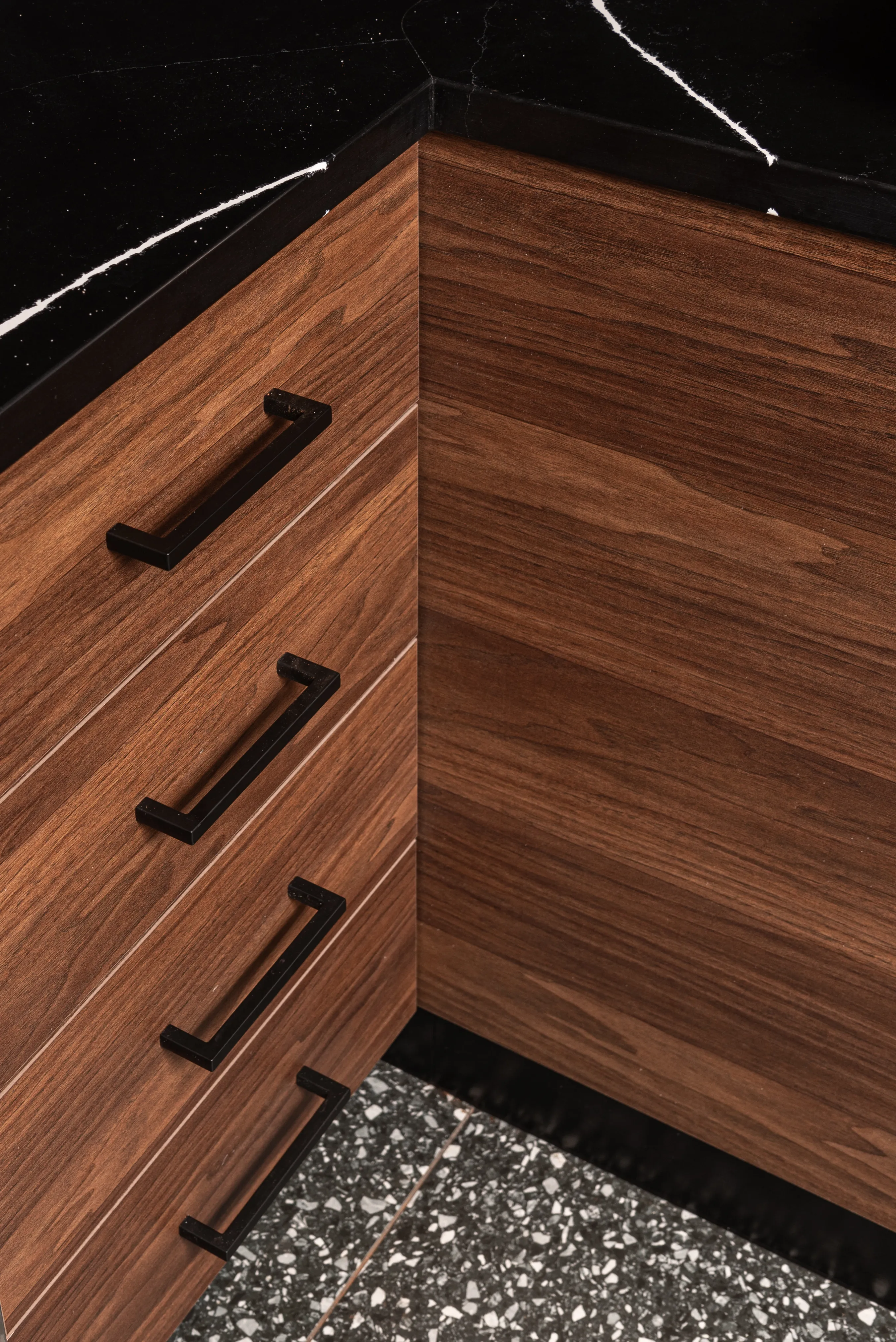 Corner of wooden kitchen cabinets with black handles and a black marble countertop above them.