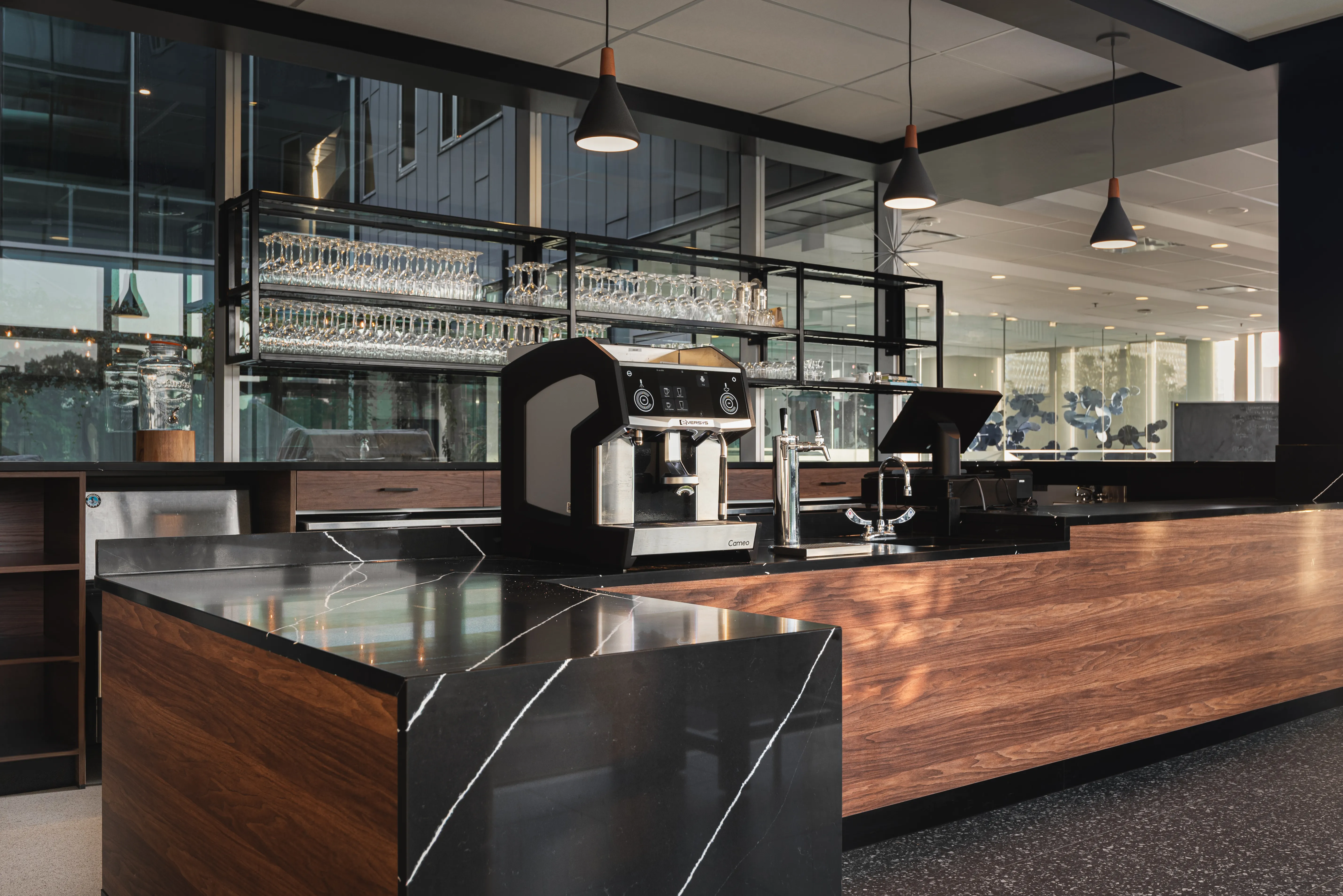 Modern coffee station with espresso machine, glassware shelves, and wood and black stone countertops.
