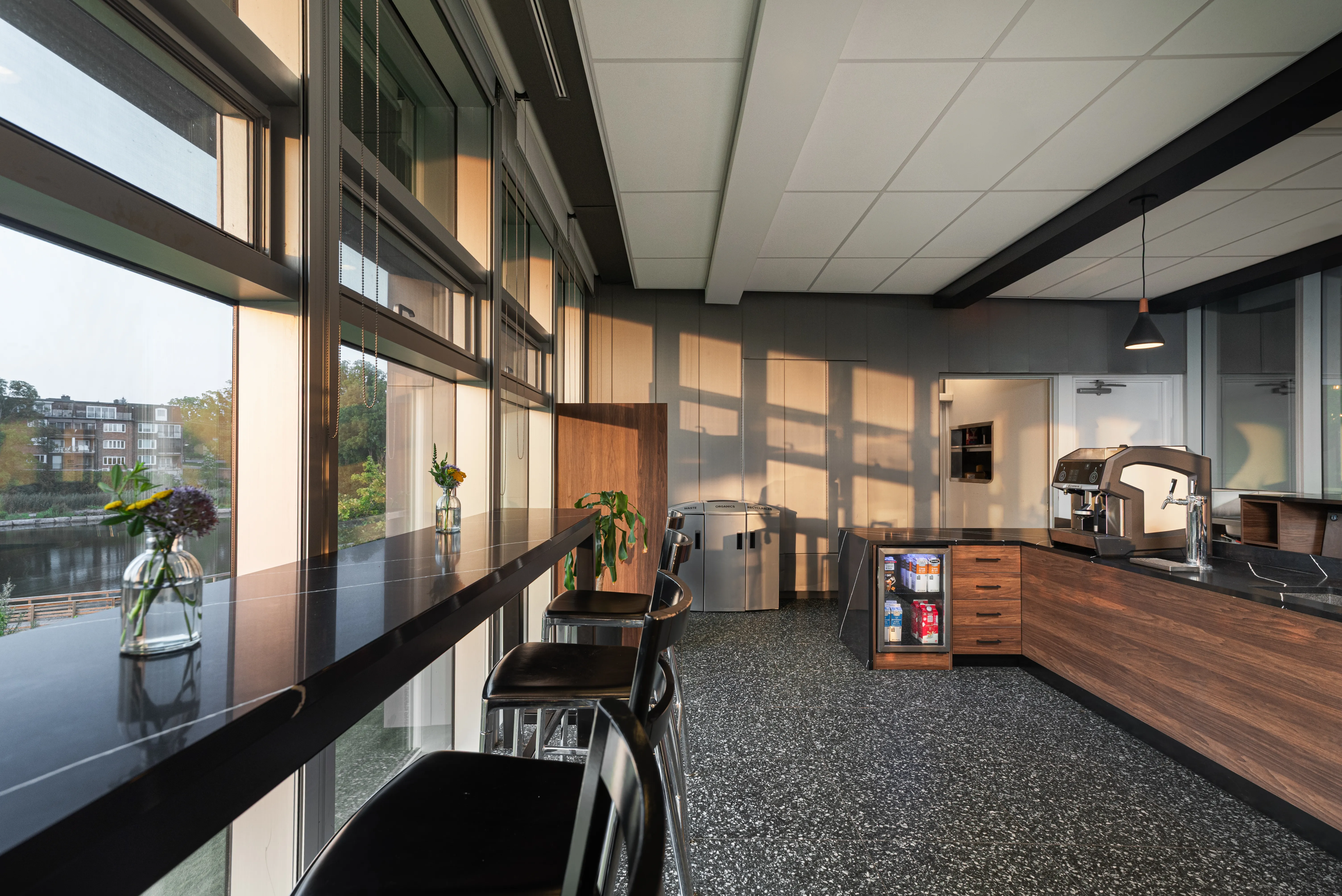 Modern kitchen with black countertop bar stools and large windows showing outdoor scenery.