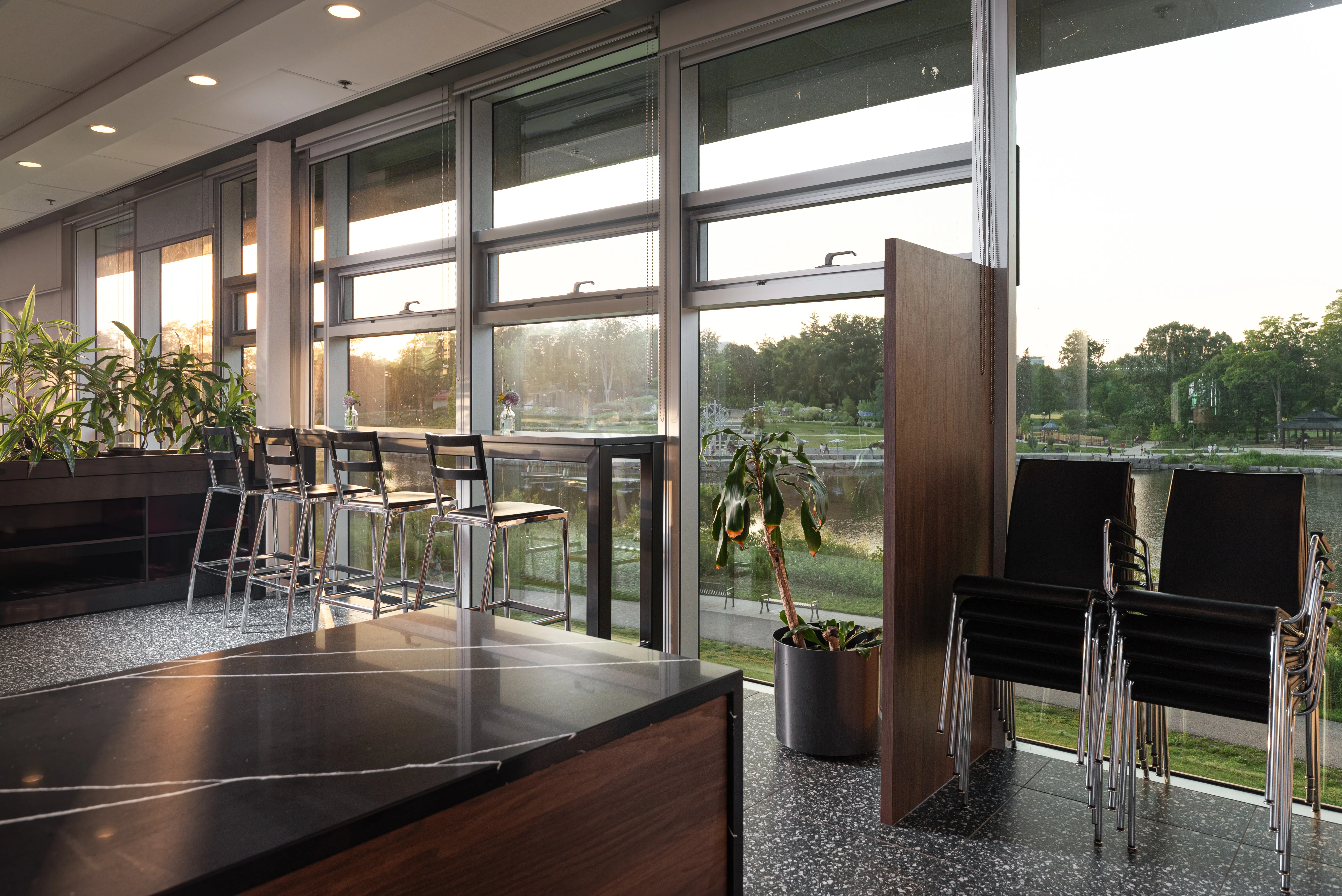 Modern room with bar stools, black chairs stacked, large windows overlooking a calm park and water.