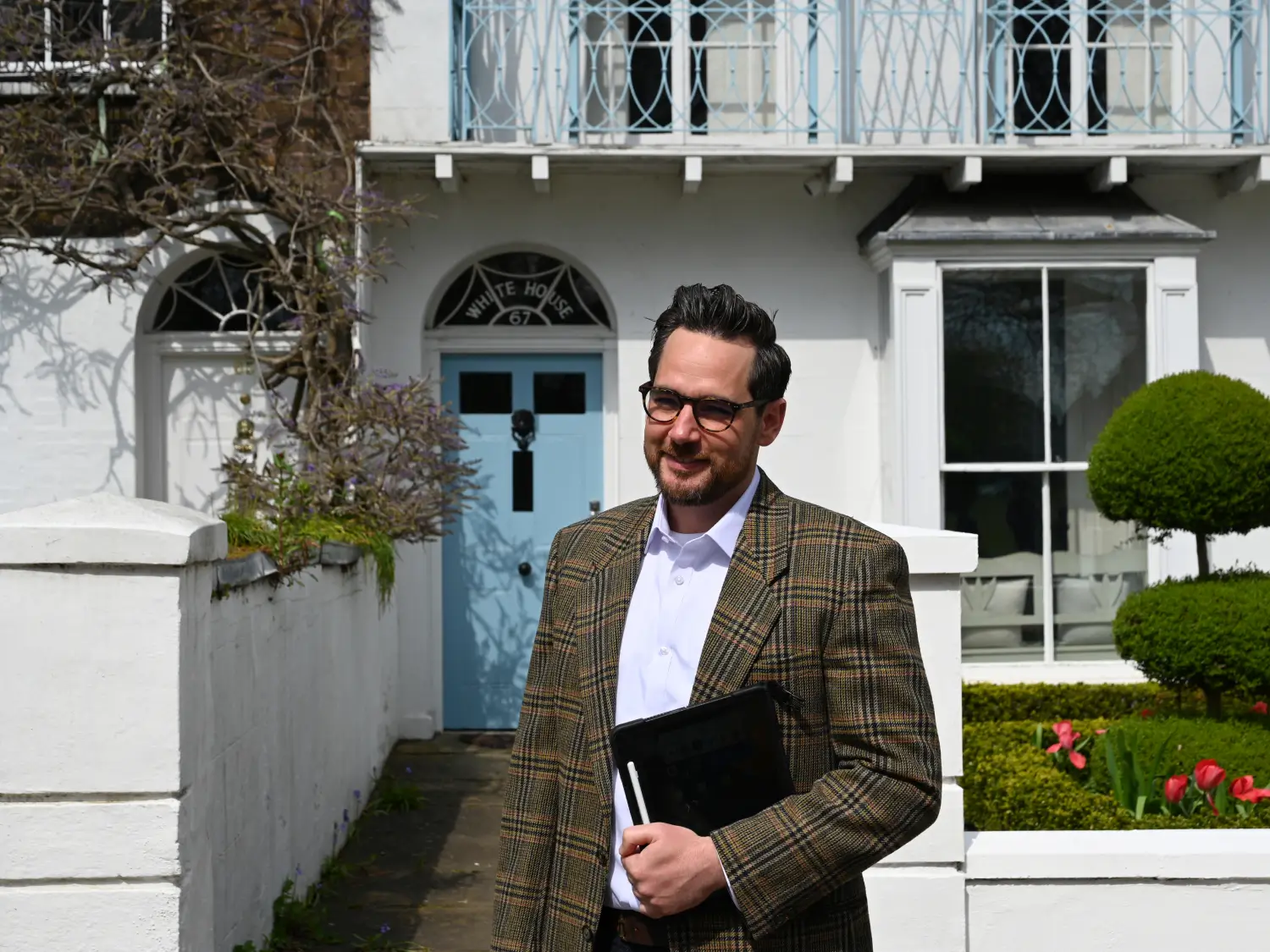 "Professional male surveyor in tweed jacket holding clipboard standing outside a white residential property"