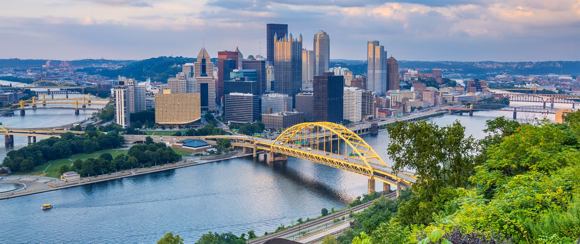 View of Pittsburgh skyline with yellow bridges crossing rivers and lush green trees in the foreground.