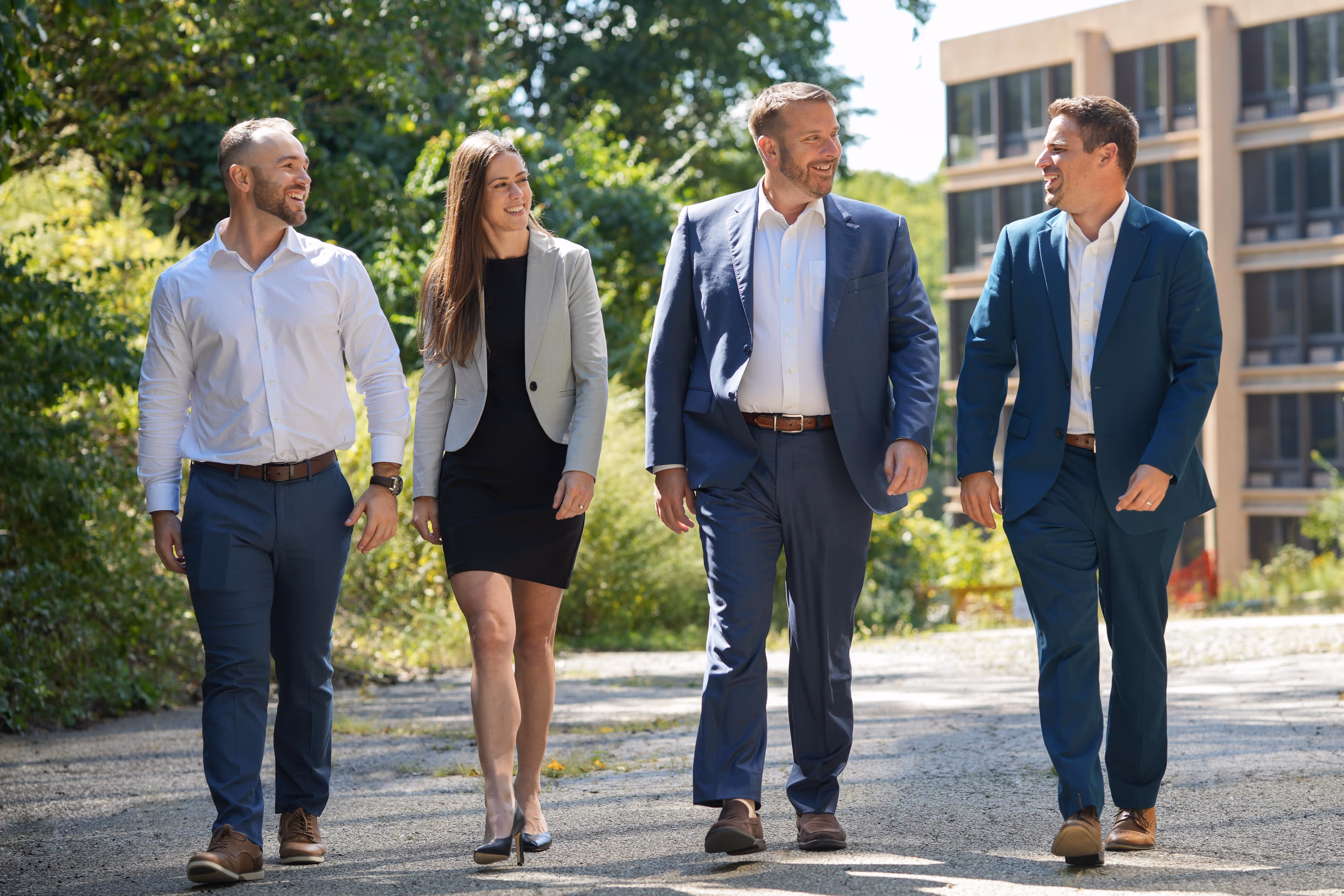 Four business professionals smiling and walking outdoors on a sunny day with greenery and a building in the background.