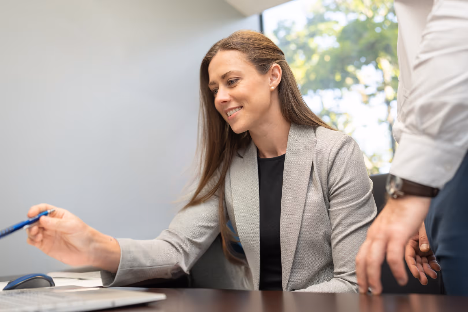 Smiling businesswoman in a gray blazer pointing at a laptop screen during a meeting.