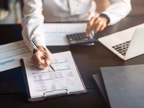 Person using a calculator and pointing at financial charts on a clipboard beside a laptop on a desk.