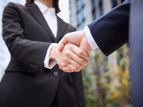Two business professionals in suits shaking hands outdoors near office buildings.