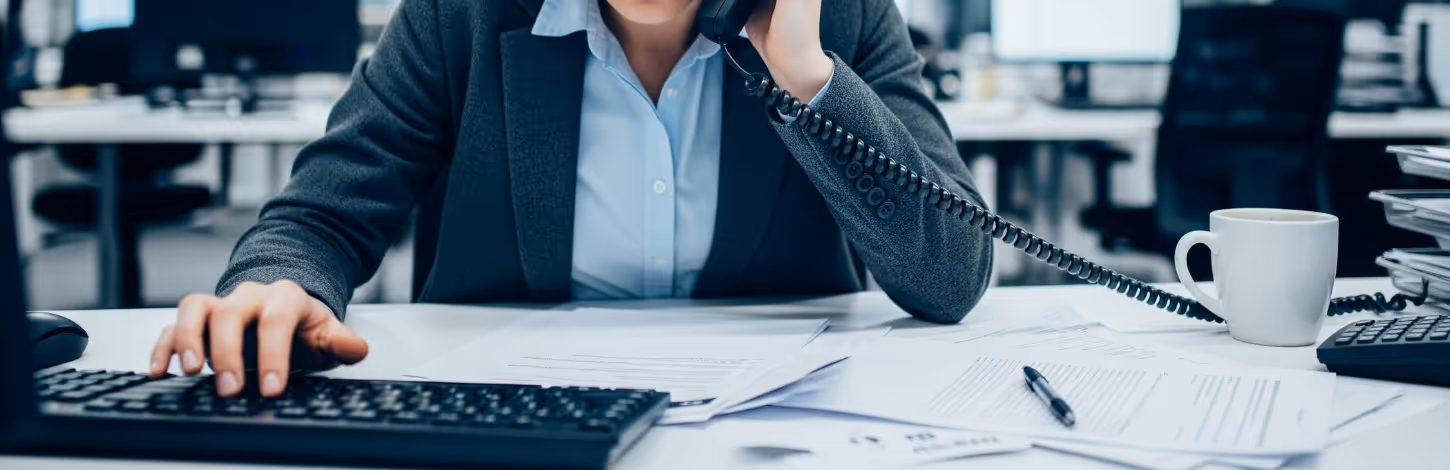 Businessperson in blazer talking on phone while working on computer at a desk with papers and coffee cup.