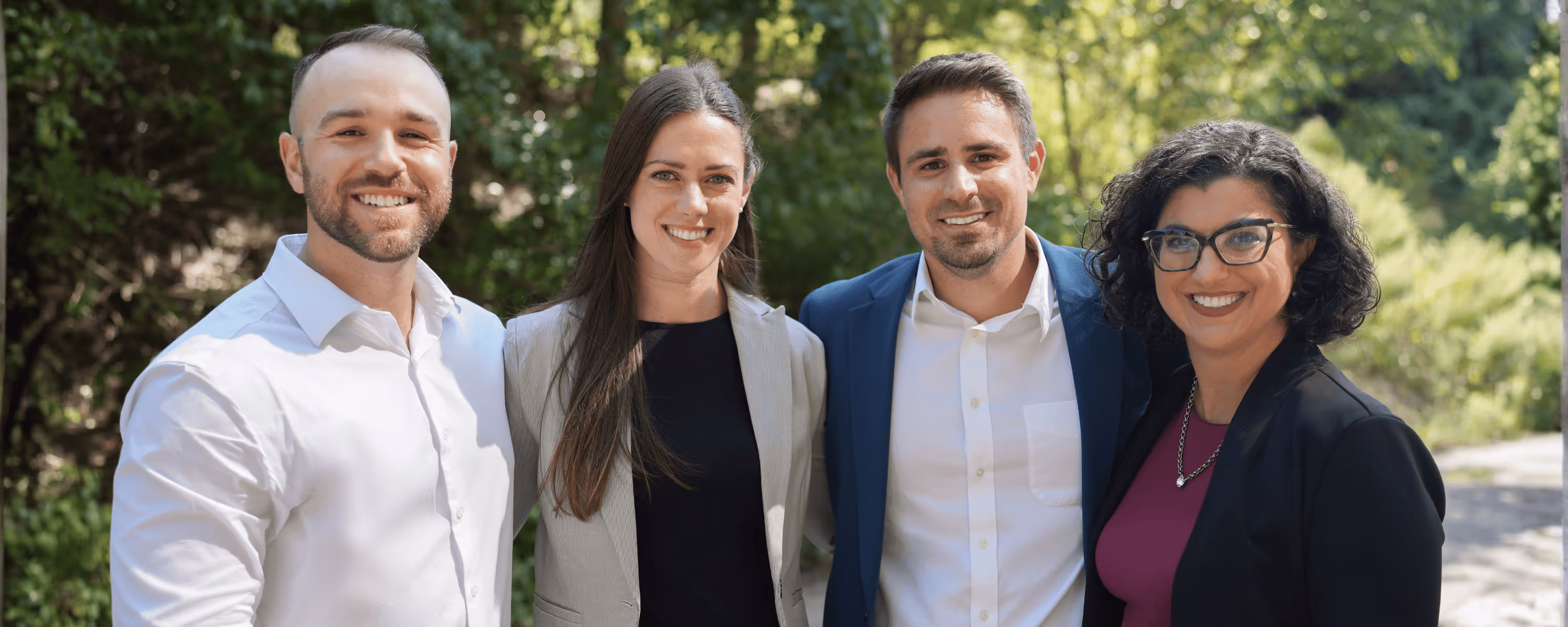Four smiling professionals standing outdoors with greenery in the background.