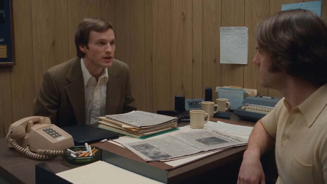 Two men talking across a wood-paneled office desk with papers, cups, and a rotary phone.