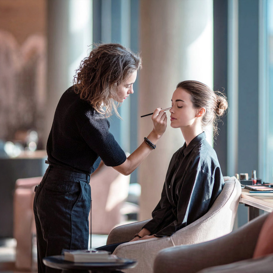 Makeup artist applying eyeshadow on a seated woman with her hair in a bun in a well-lit room.