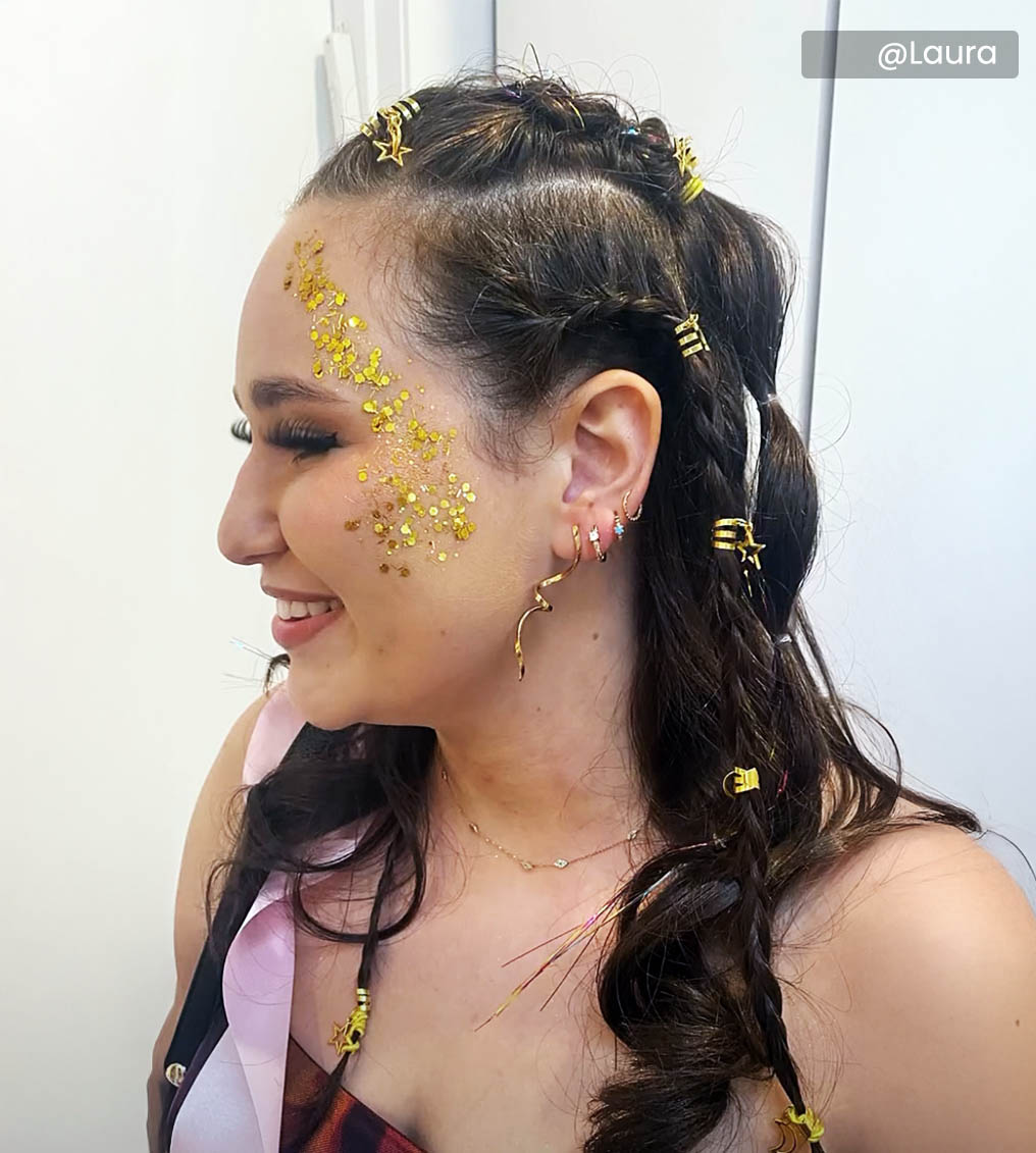 Young woman with vibrant rainbow eye makeup and glossy lips holding a cup filled with paintbrushes, wearing a yellow top.
