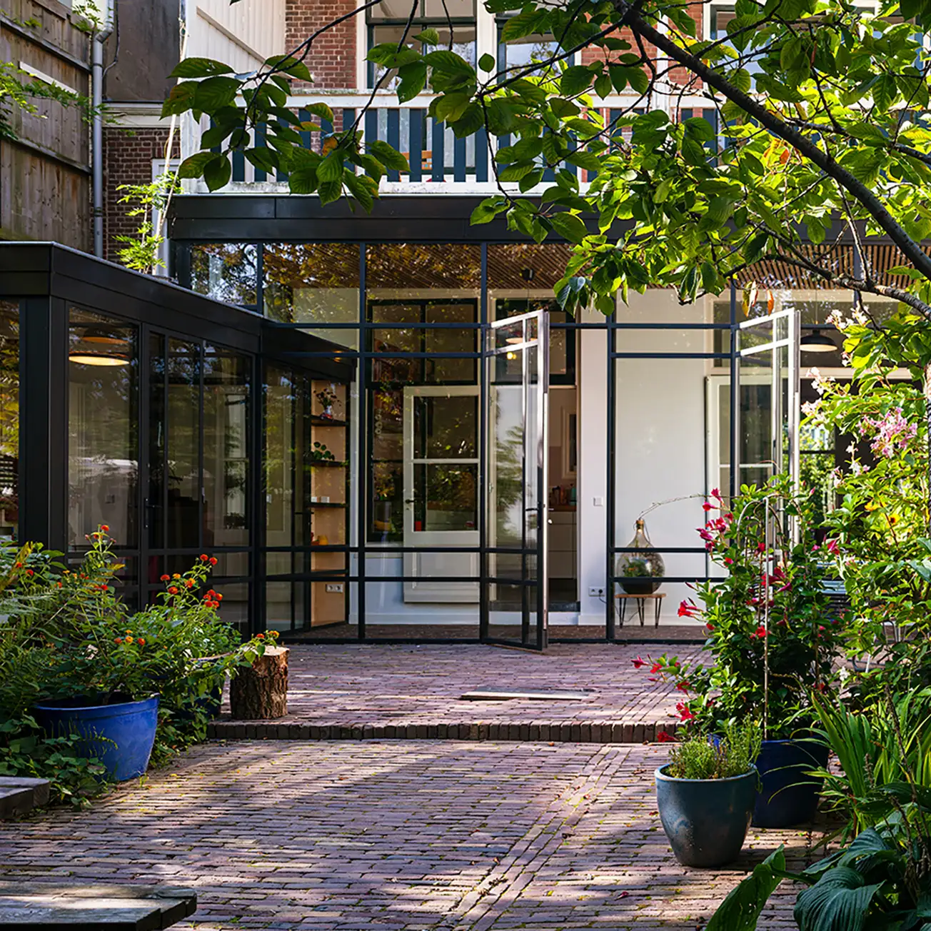Modern house extension with large glass doors opening to a brick patio surrounded by green plants and potted flowers.
