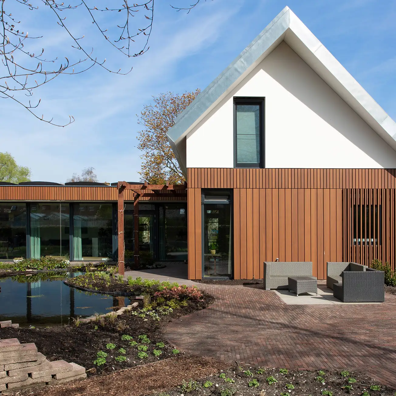 Modern house with white upper gable and vertical wooden paneling, a small pond with surrounding garden, and outdoor patio furniture.