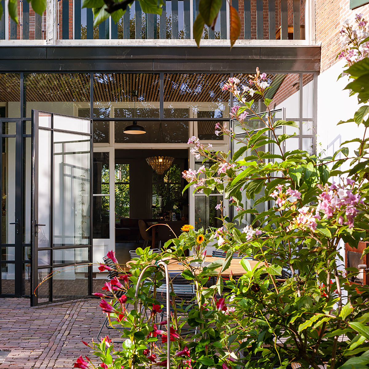 Open glass doors leading to a patio with a wooden table surrounded by chairs and colorful flowering plants in front.