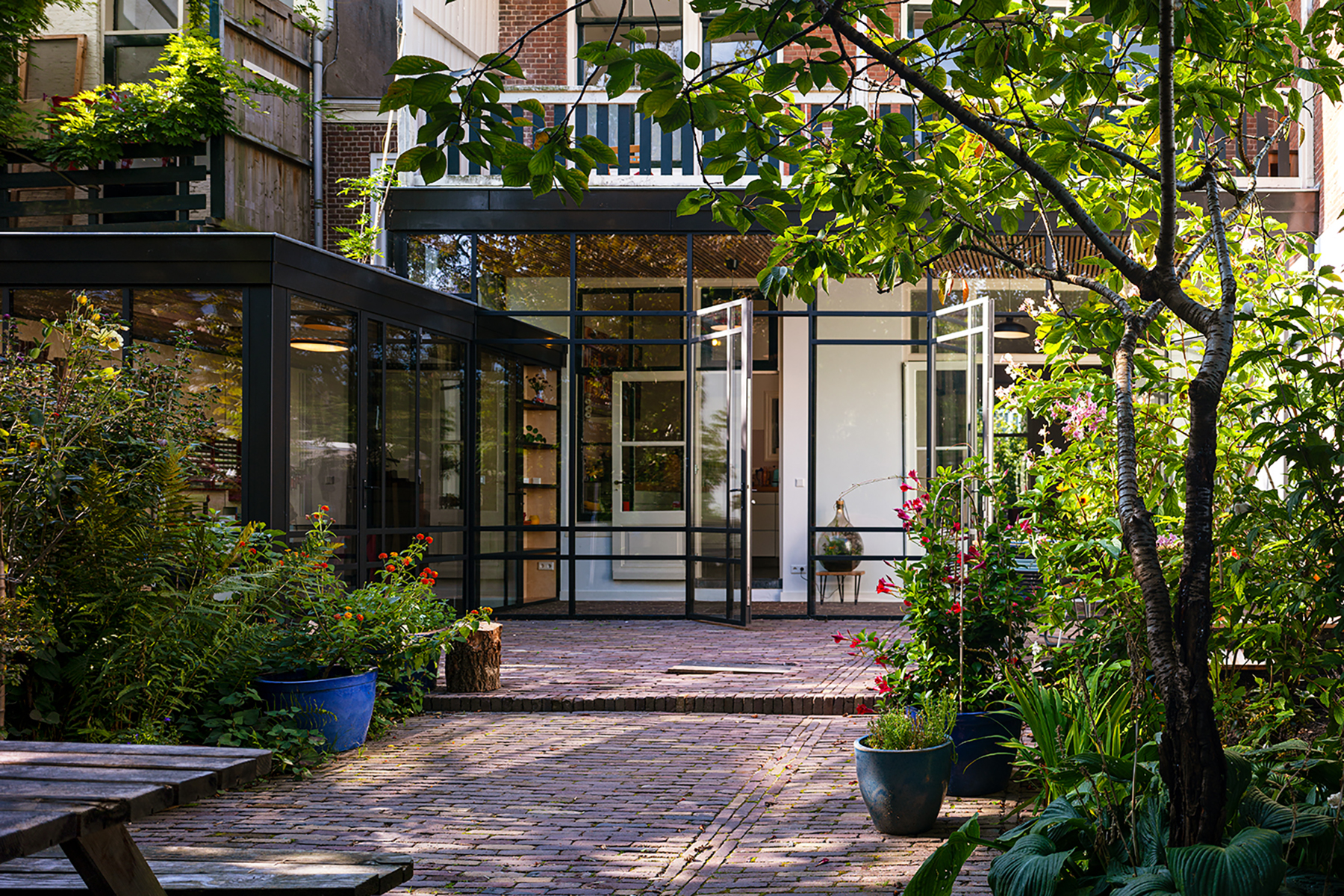Sunlit brick patio garden with various potted plants and a glass wall entrance to a modern building.