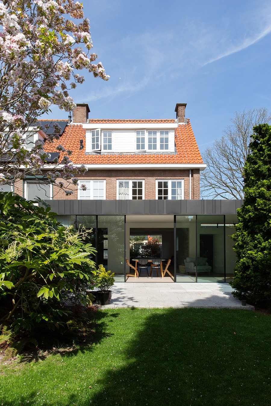Backyard view of a two-story brick house with a red tile roof and large modern glass extension opening to a green lawn.