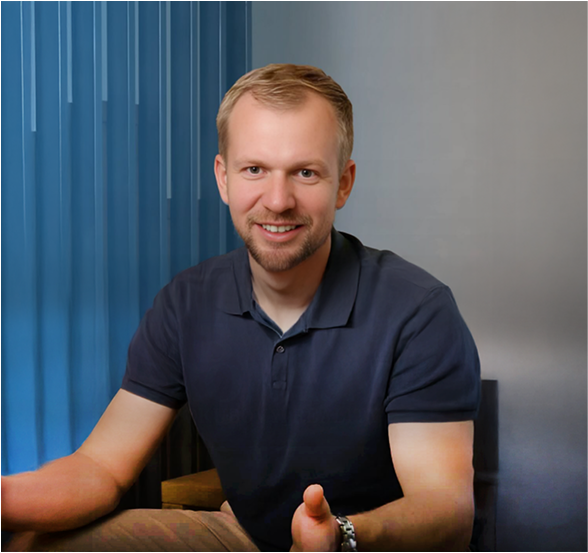 Muž s krátkými světlými vlasy a strništěm v tmavě modrém polo tričku sedí a usmívá se směrem k divákovi.