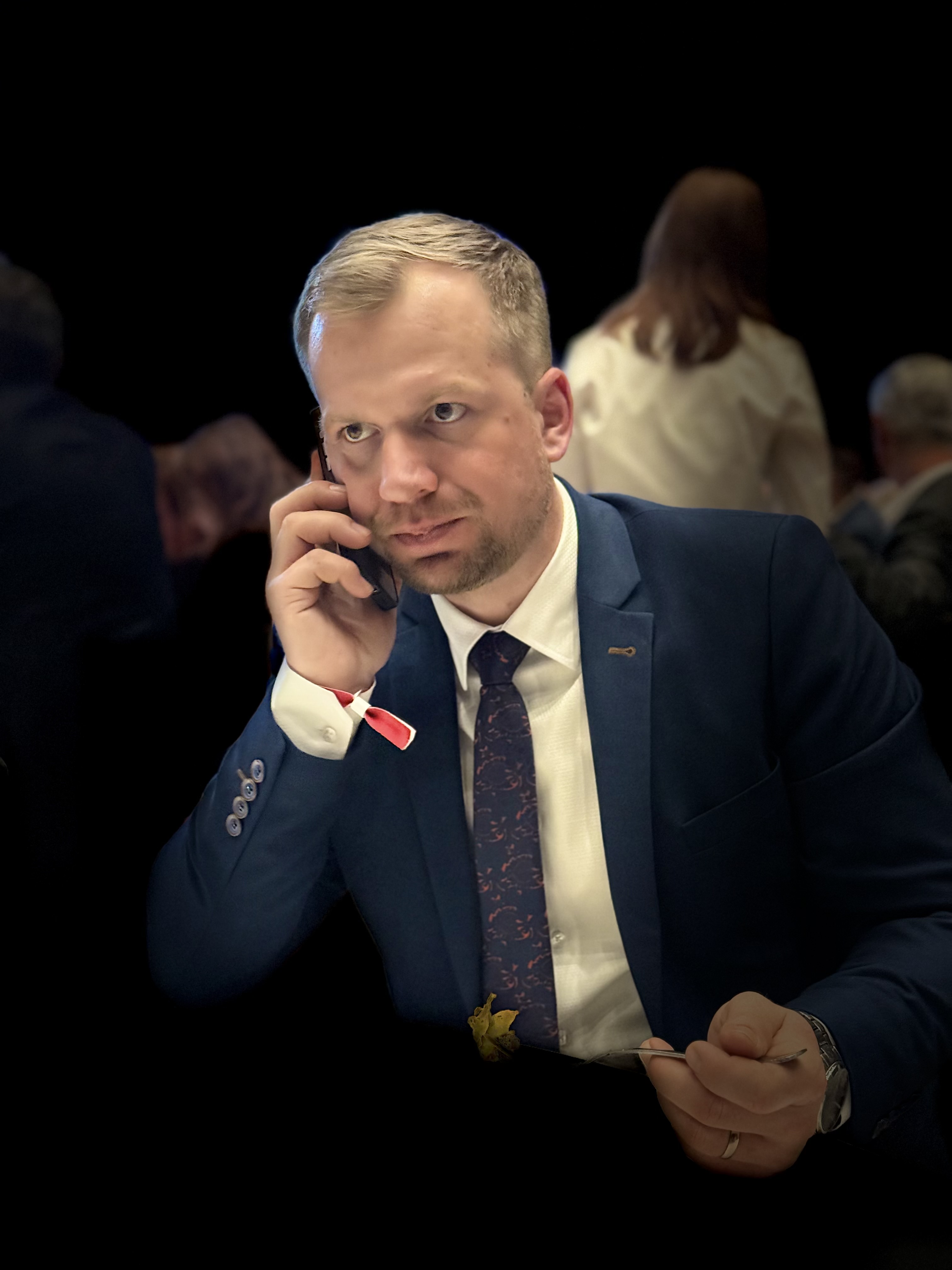 Muž ve tmavě modrém obleku mluví na mobilní telefon a drží vidličku s jídlem.