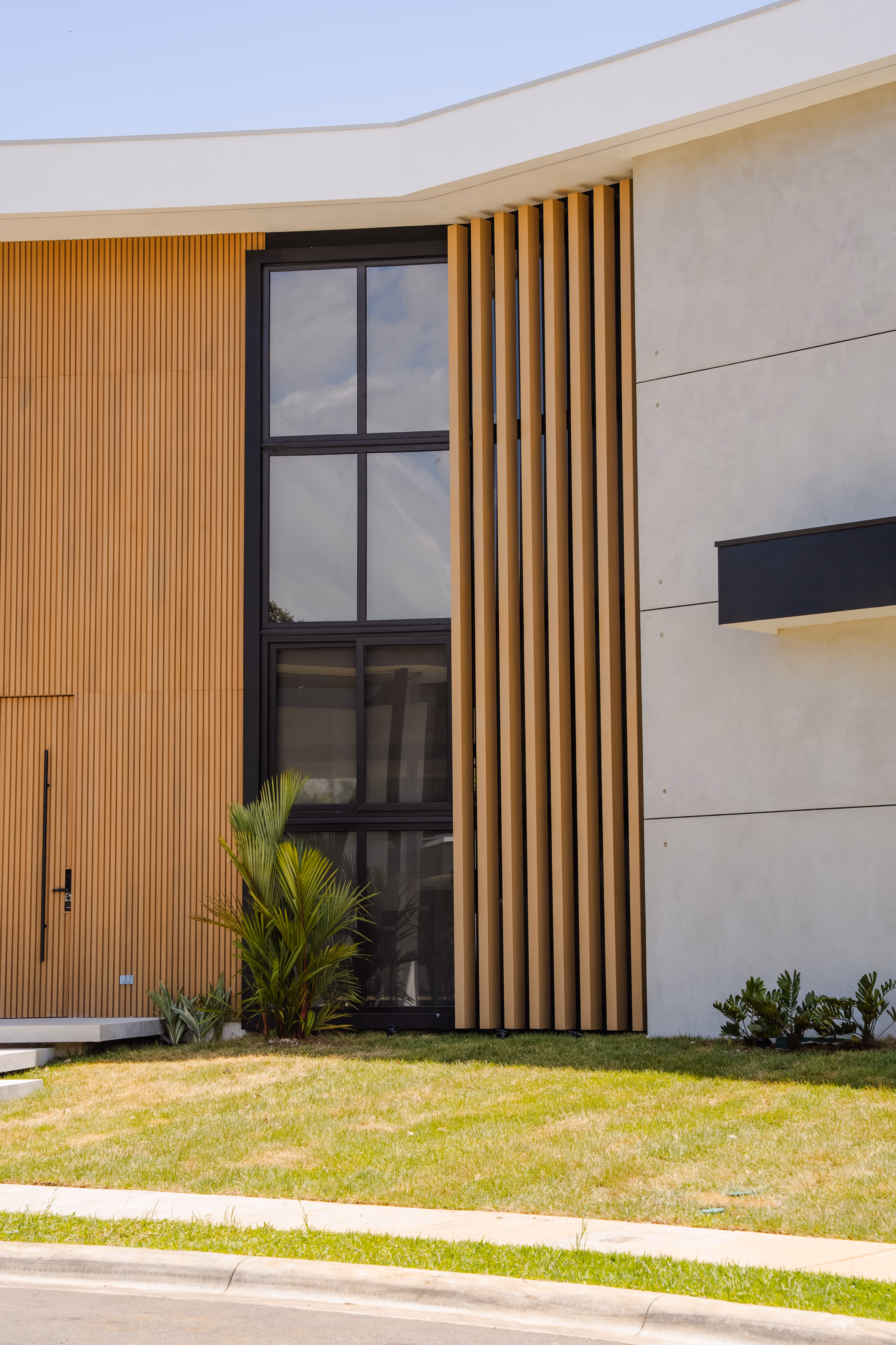 Fachada moderna de casa con paredes beige y paneles verticales de madera, ventana grande de vidrio en pvc negro y césped verde al frente.